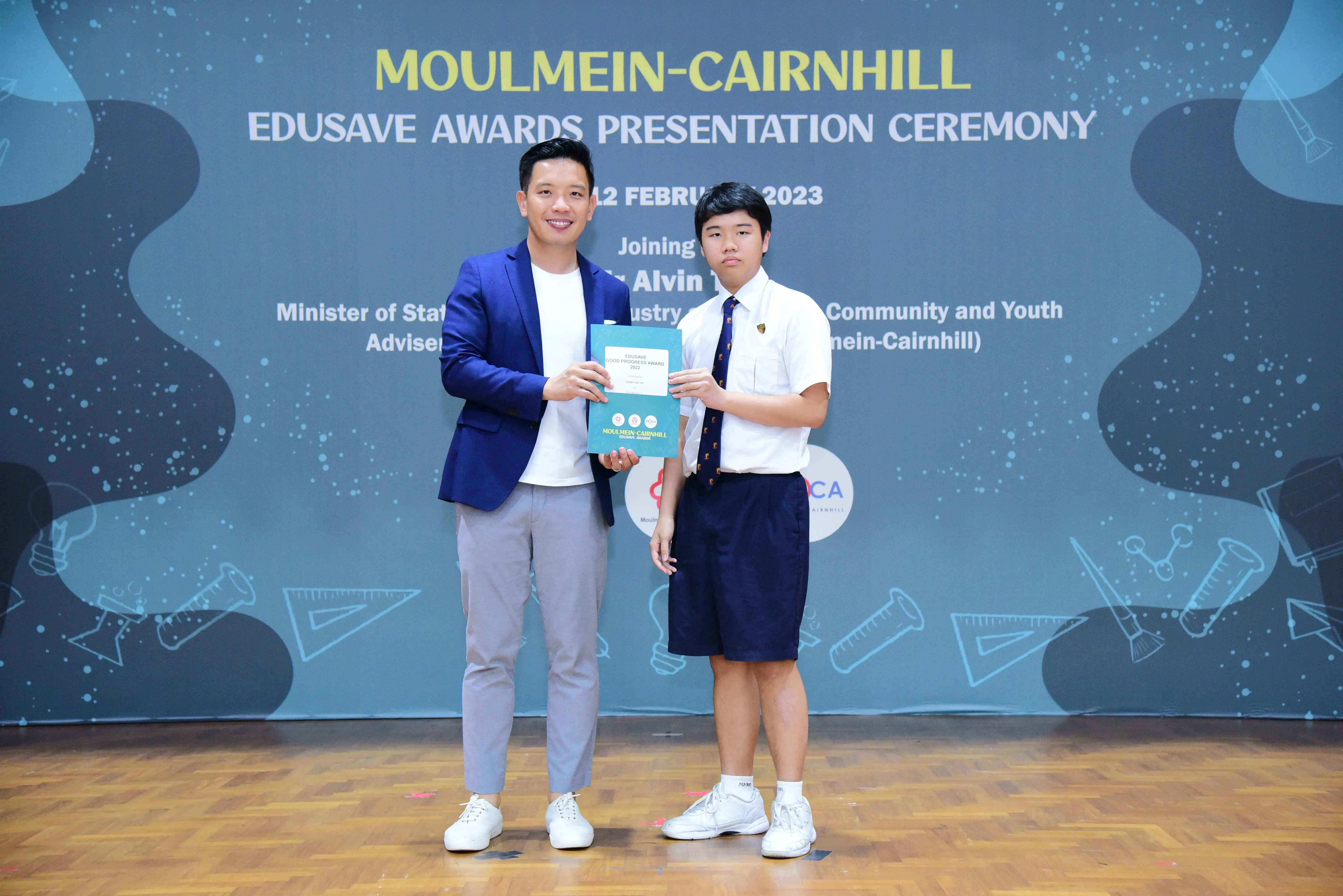 An adult in a blue blazer presents an award certificate to a uniformed student at the Moulmein-Cairnhill Edusave Awards Ceremony 2023.