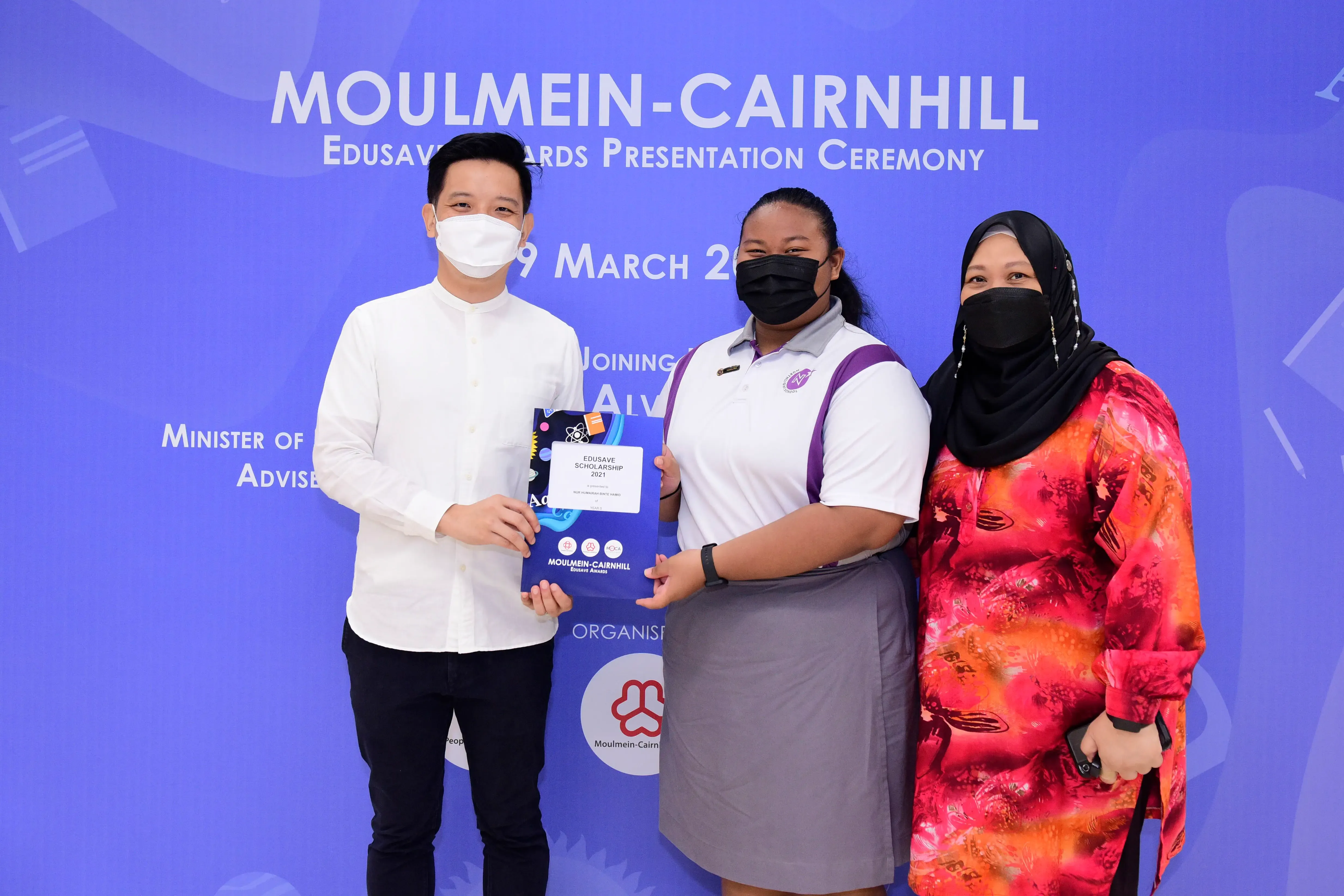 Three masked people pose at the Moulmein-Cairnhill Edusave Awards Presentation Ceremony, holding a scholarship certificate.
