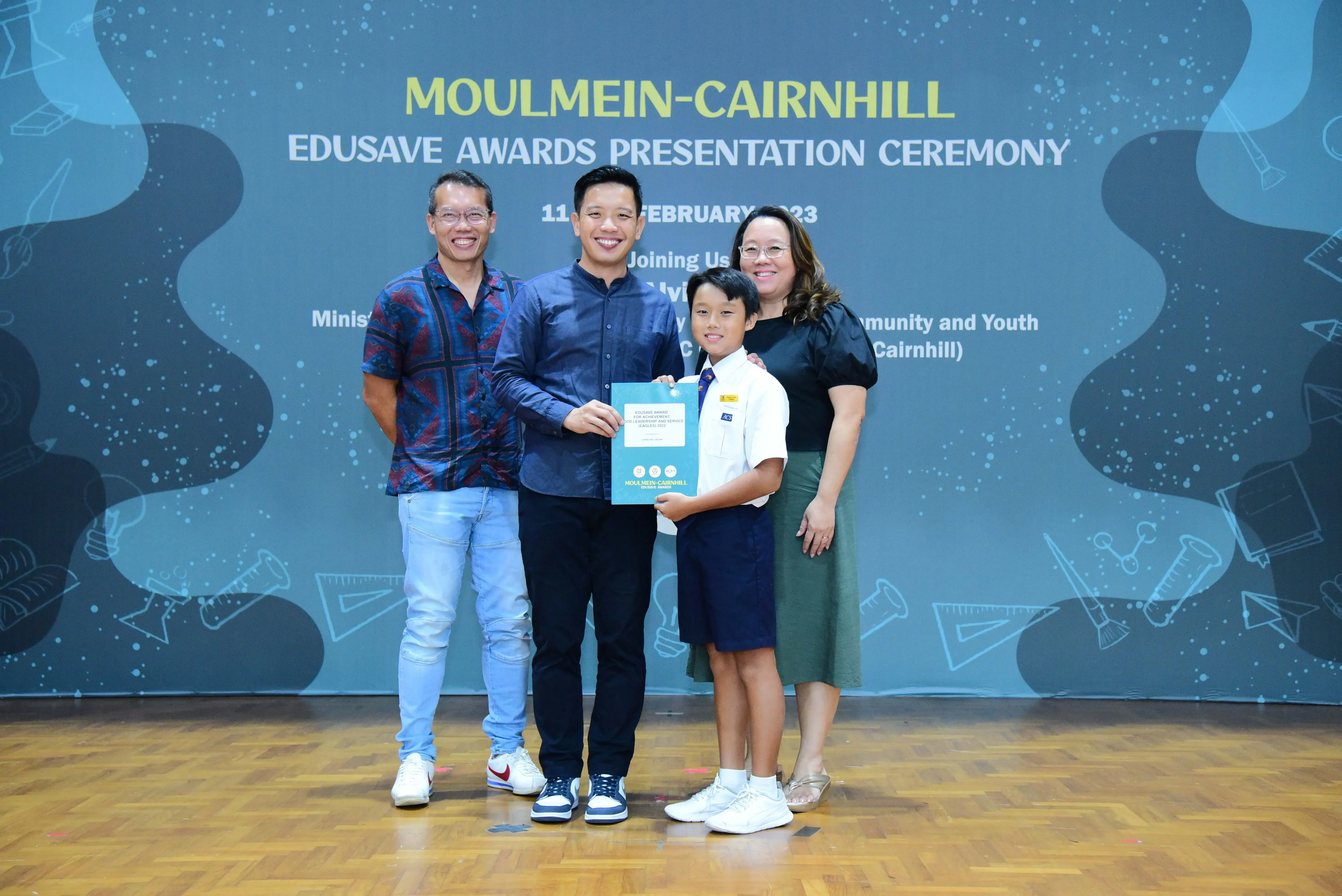 Three adults and a boy in school uniform hold a certificate at the Moulmein-Cairnhill Edusave Awards Ceremony 2023.