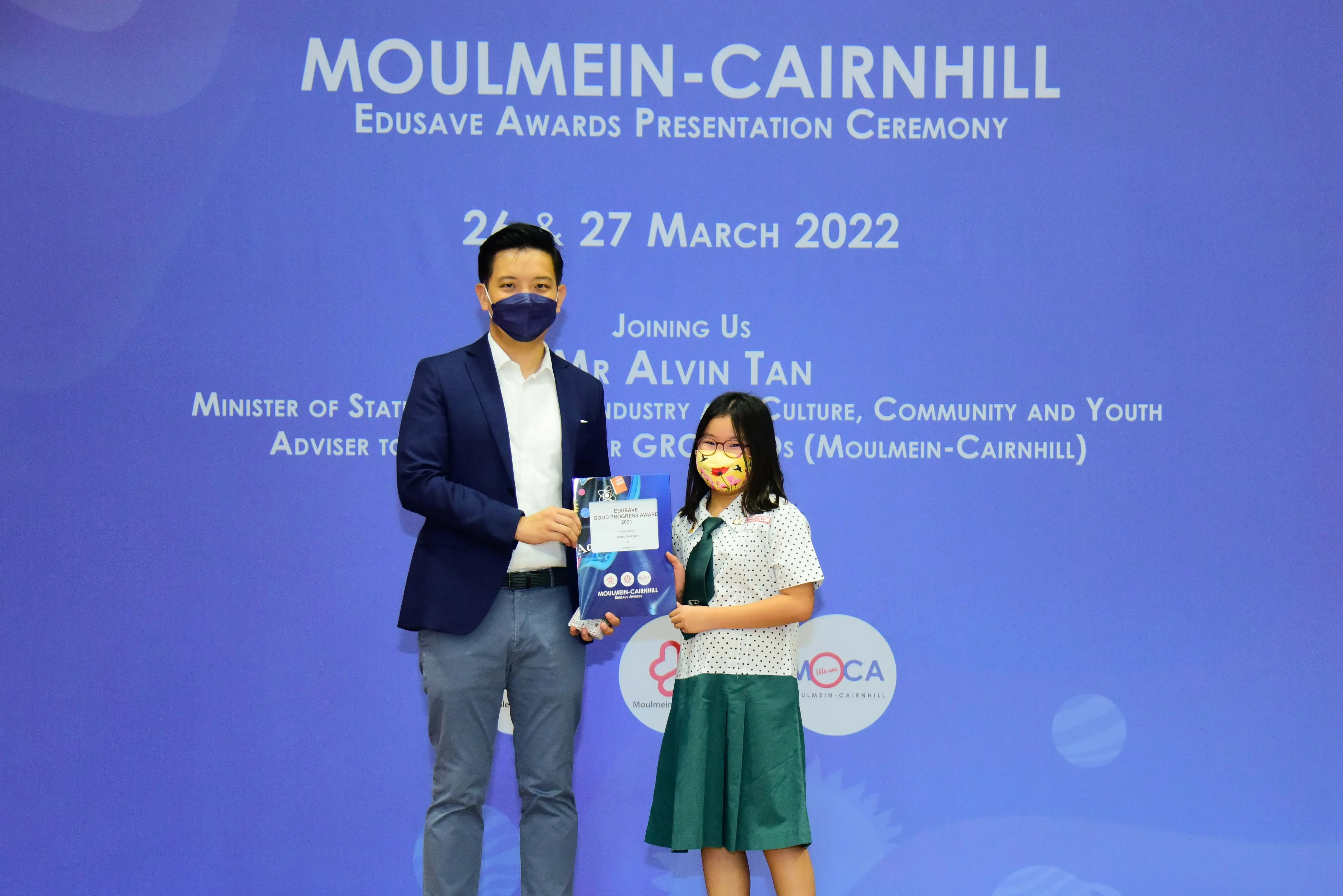 A man in a suit and a young girl in school uniform hold an award at the Moulmein-Cairnhill Edusave ceremony, March 2022.