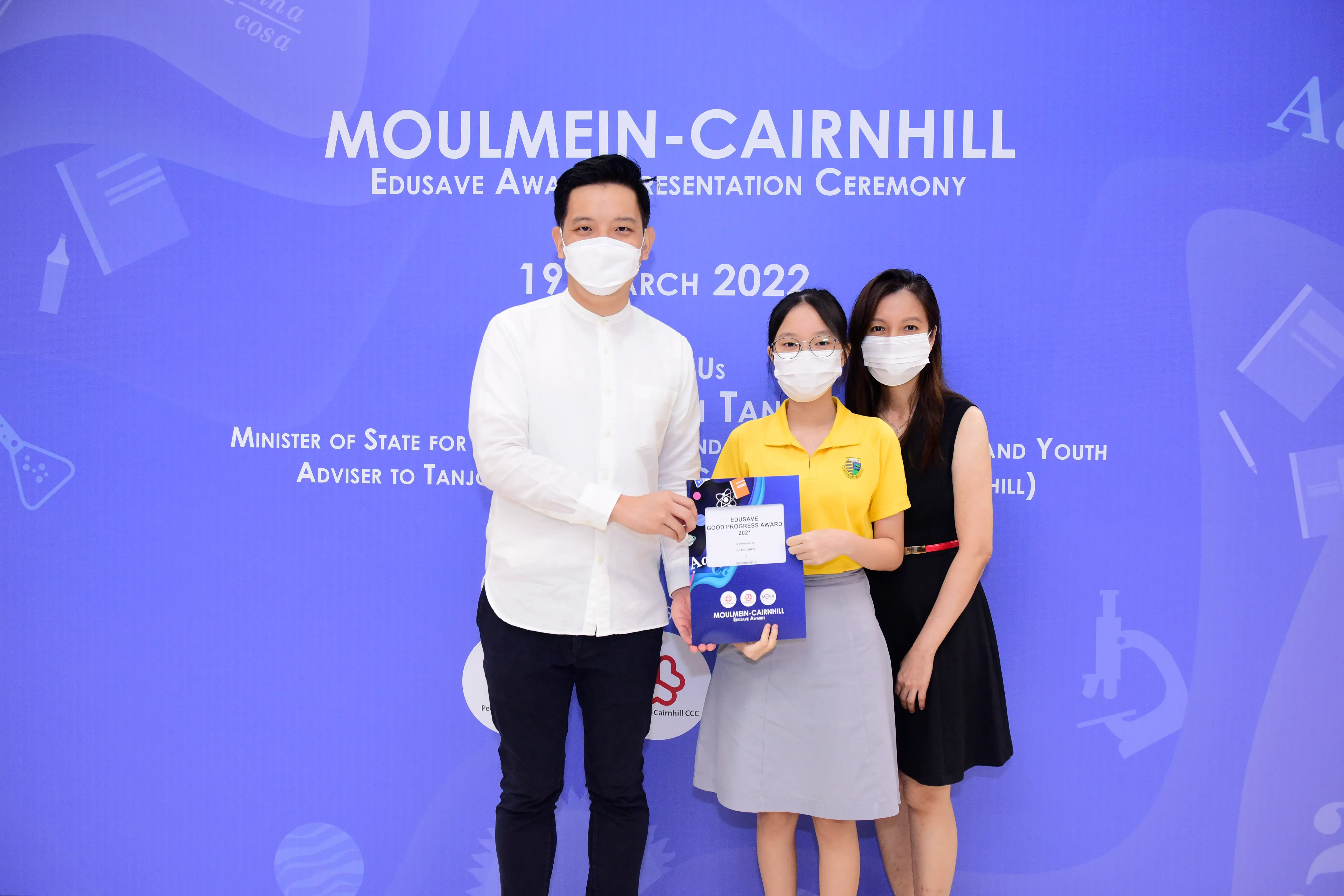 Three masked people pose at the Moulmein-Cairnhill Edusave Award Presentation Ceremony, with a girl holding an award certificate.