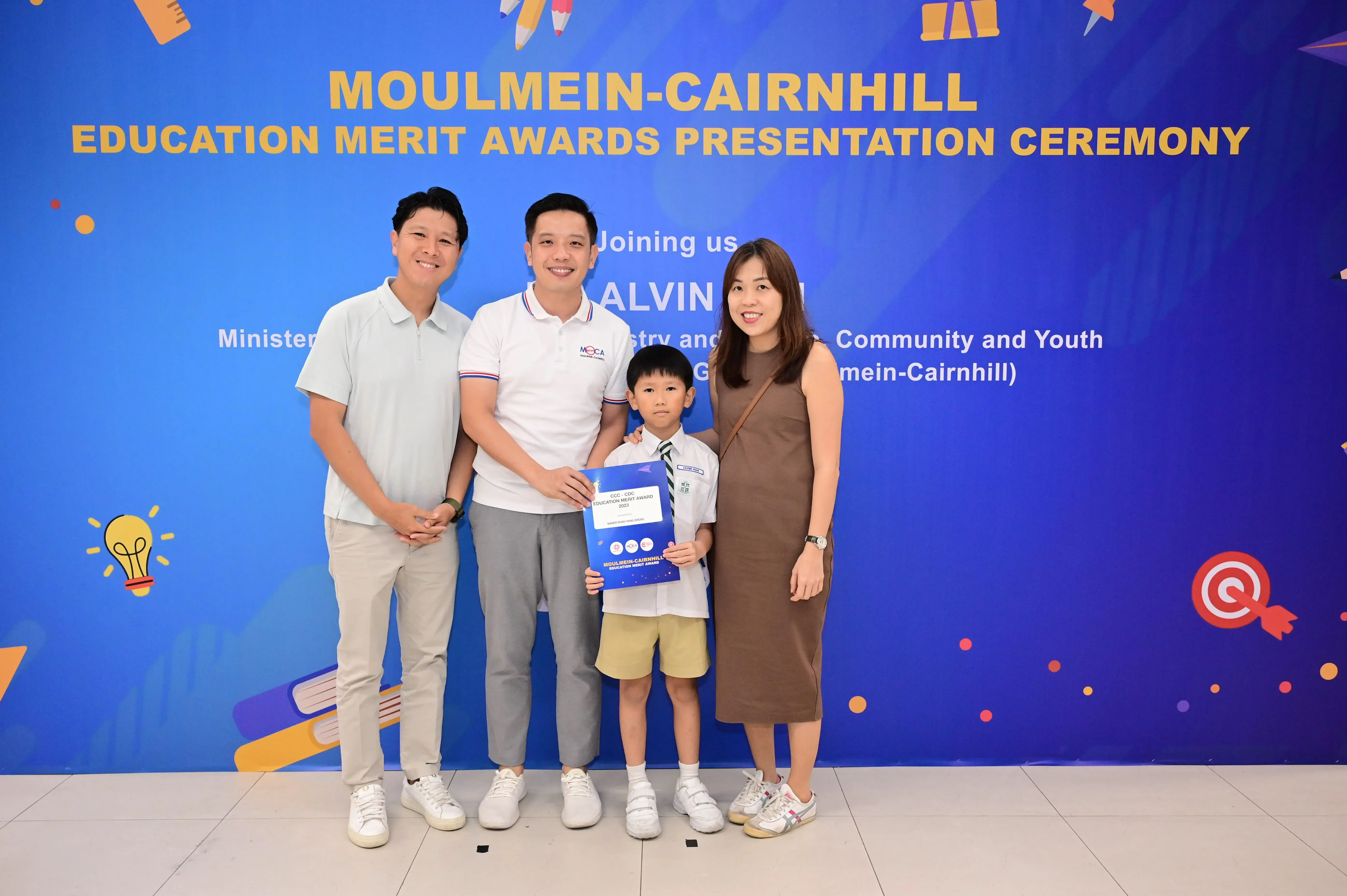 Three adults and a young boy holding an award certificate pose in front of a Moulmein-Cairnhill Education Merit Awards banner.