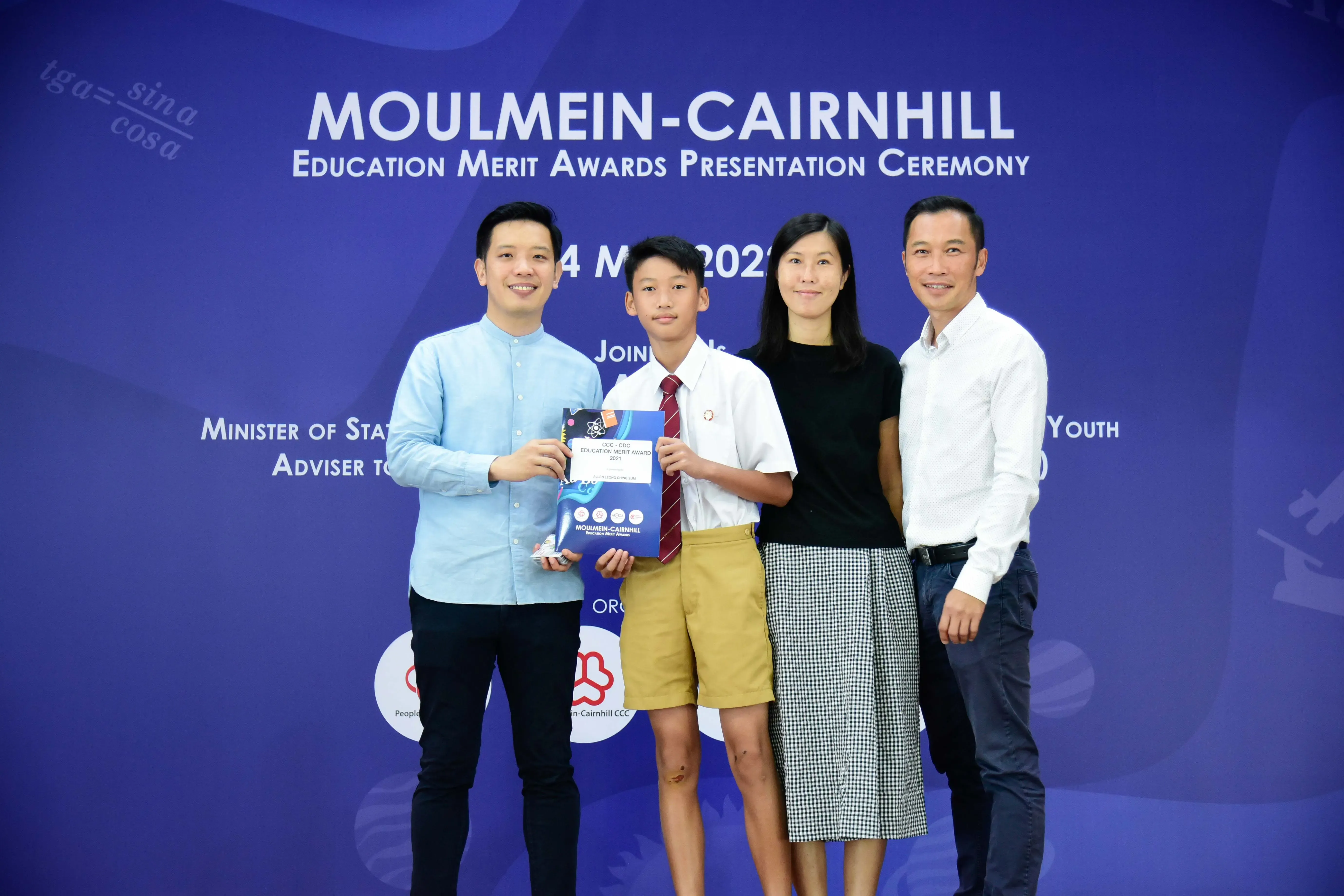 A young student holds an award certificate with three adults at the Moulmein-Cairnhill Education Merit Awards Ceremony.