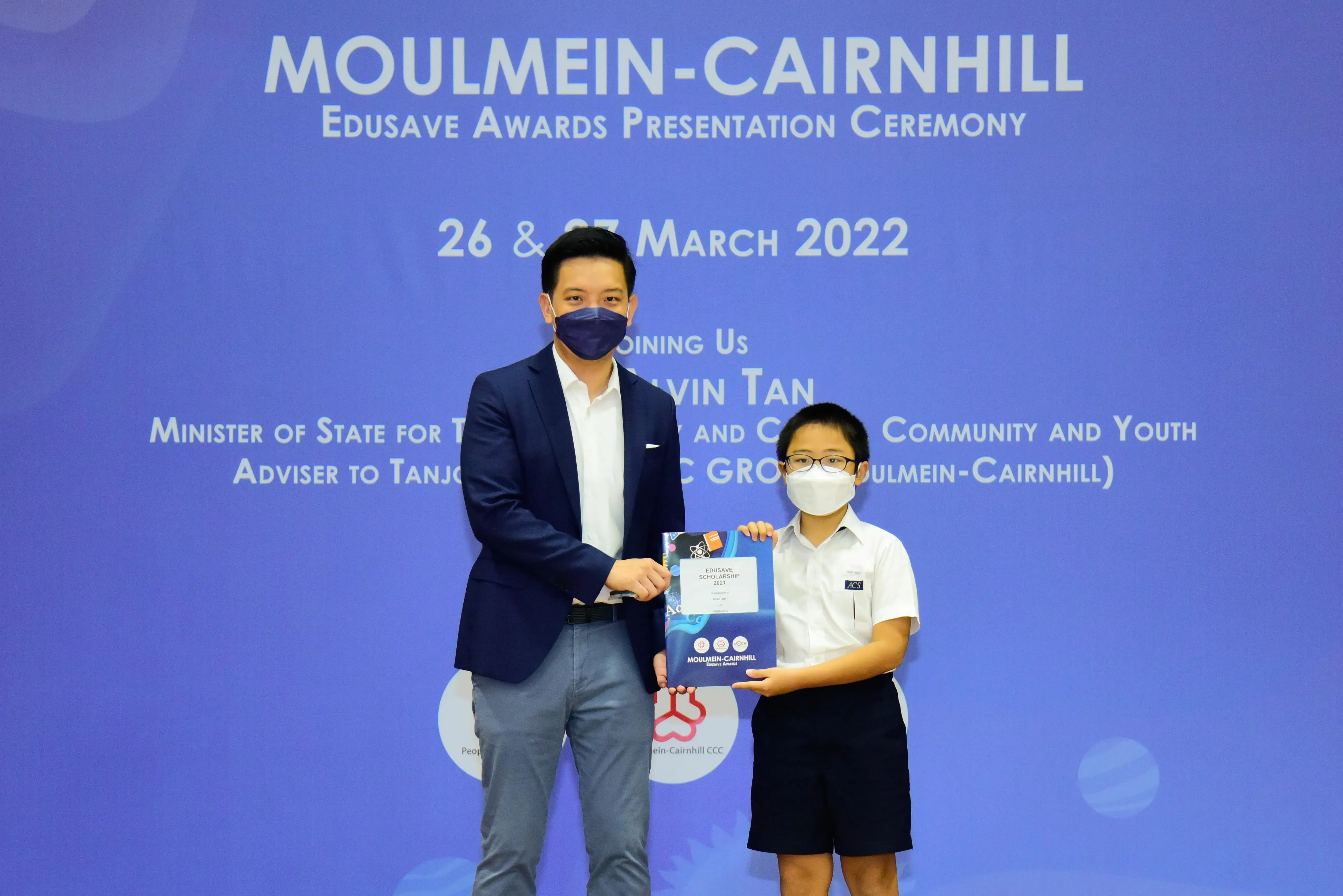 An adult and a child in masks hold an Edusave Scholarship certificate at the Moulmein-Cairnhill Awards Ceremony, March 2022.