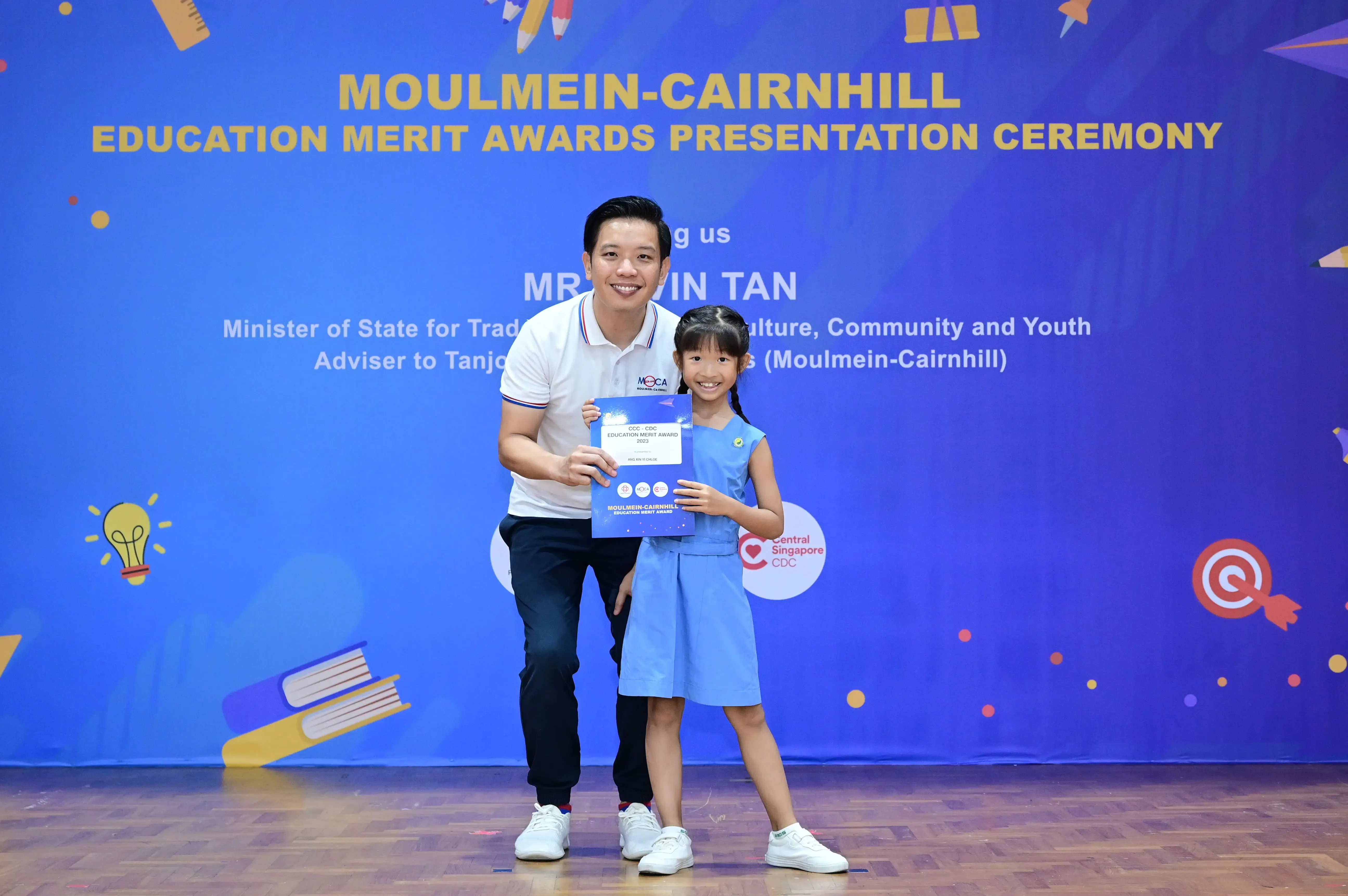 A man and a young girl hold a certificate in front of a Moulmein-Cairnhill Education Merit Awards banner.