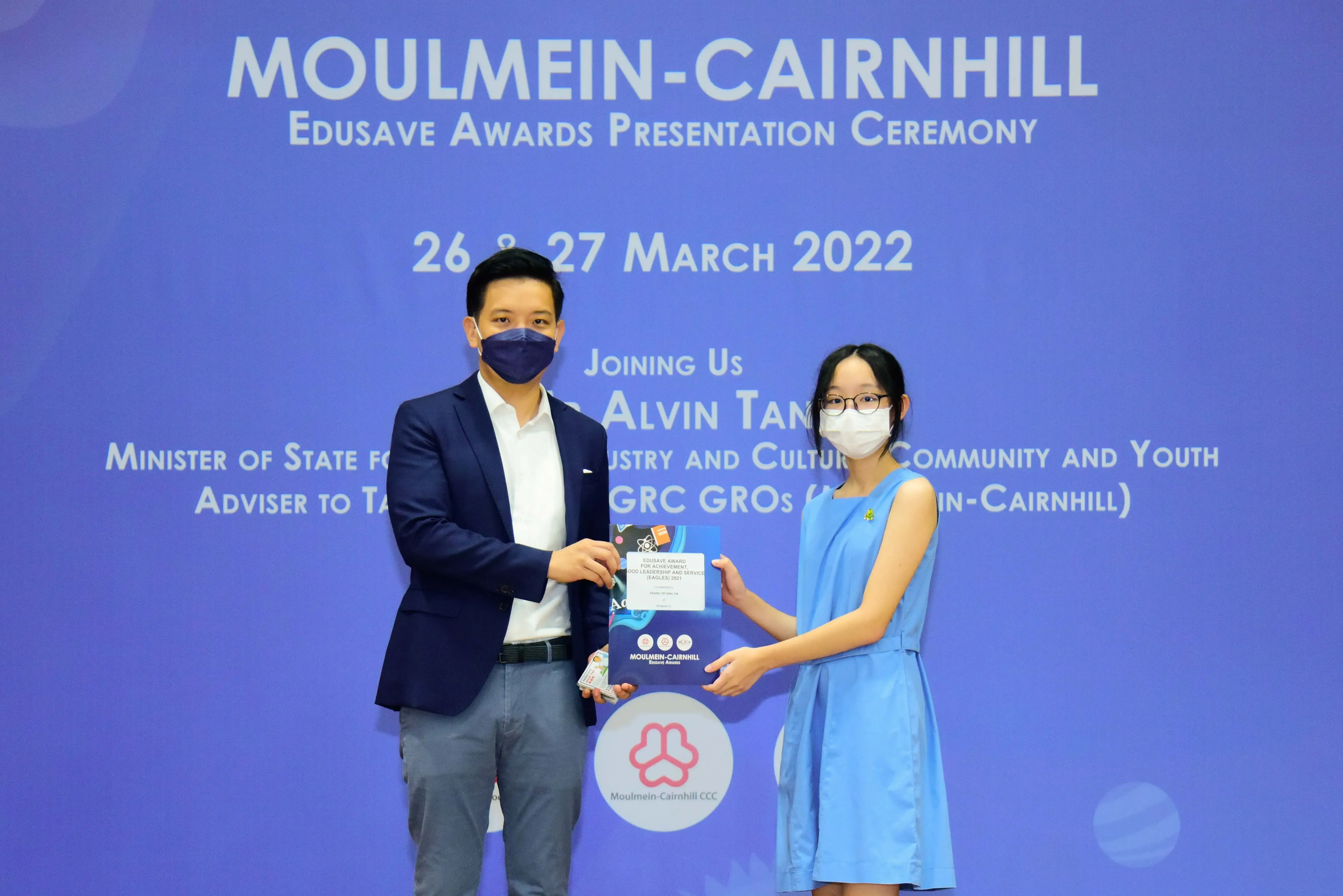Two masked people hold an award folder at the Moulmein-Cairnhill Edusave Awards Presentation Ceremony, March 2022.