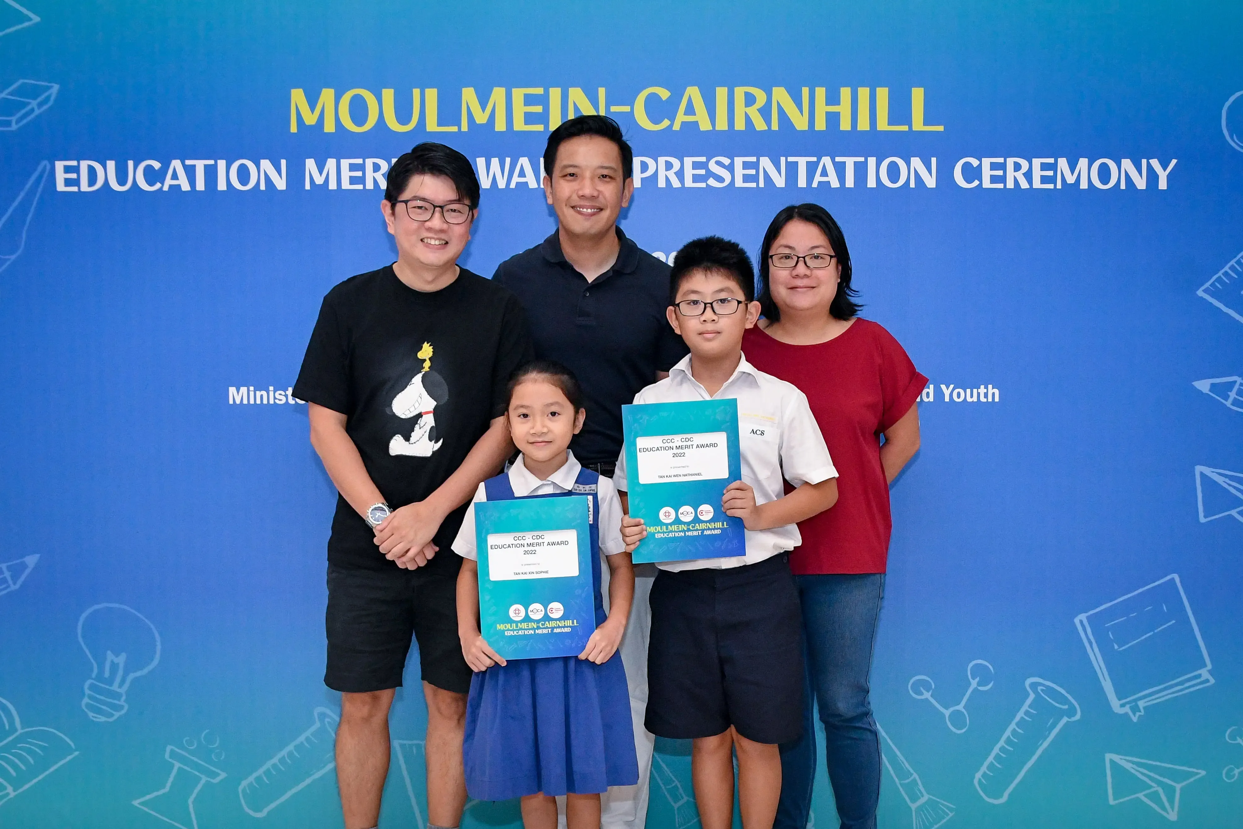 Five people pose in front of a Moulmein-Cairnhill Education Merit Award Presentation Ceremony banner, two children holding award folders.