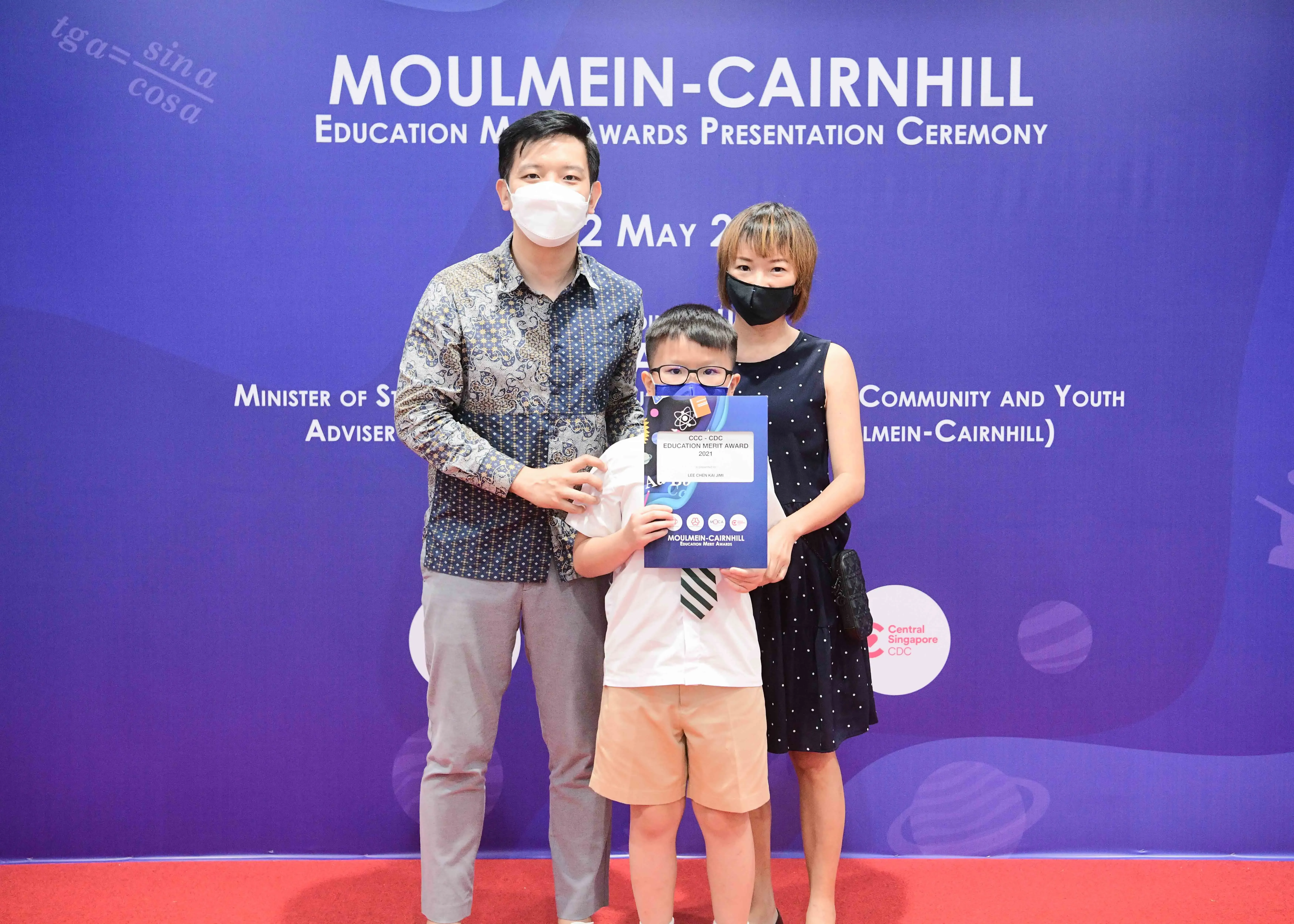 Two masked adults and a young boy holding an award certificate pose at the Moulmein-Cairnhill Education Merit Awards ceremony.