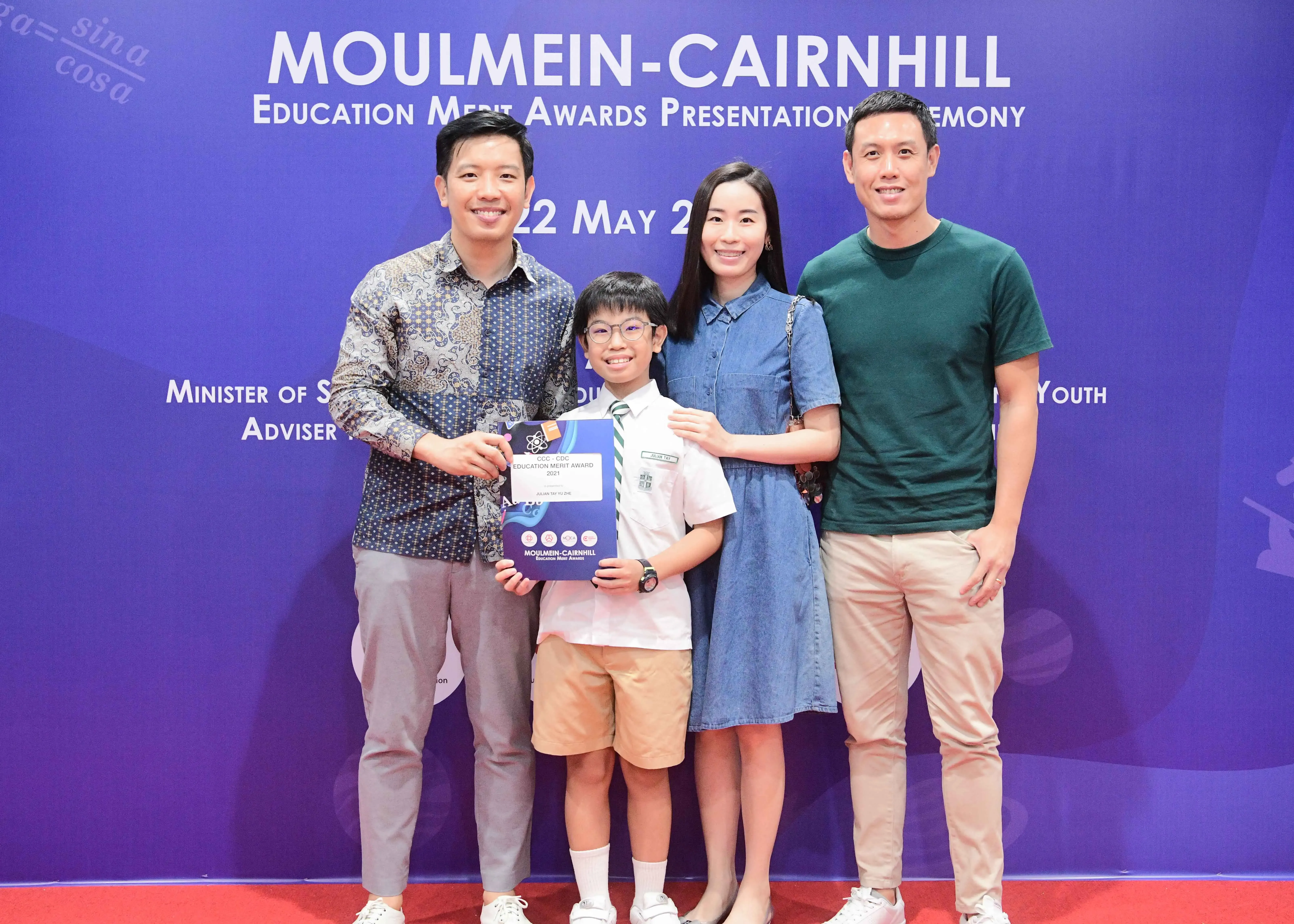 Four people pose smiling at the Moulmein-Cairnhill Education Merit Awards, with a child holding an award certificate.