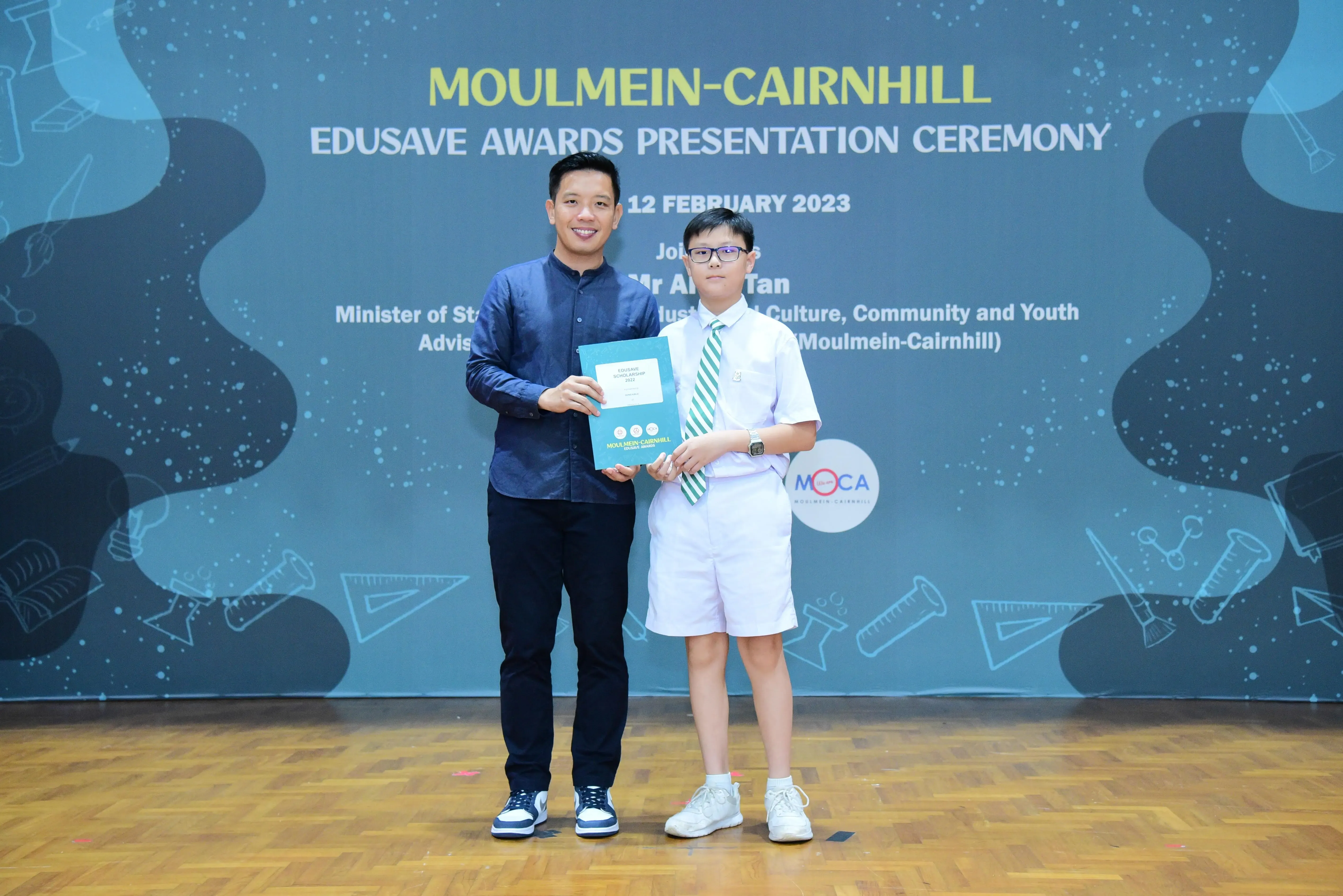 A man and a young student in uniform hold an award booklet at the Moulmein-Cairnhill Edusave Awards Ceremony 2023.