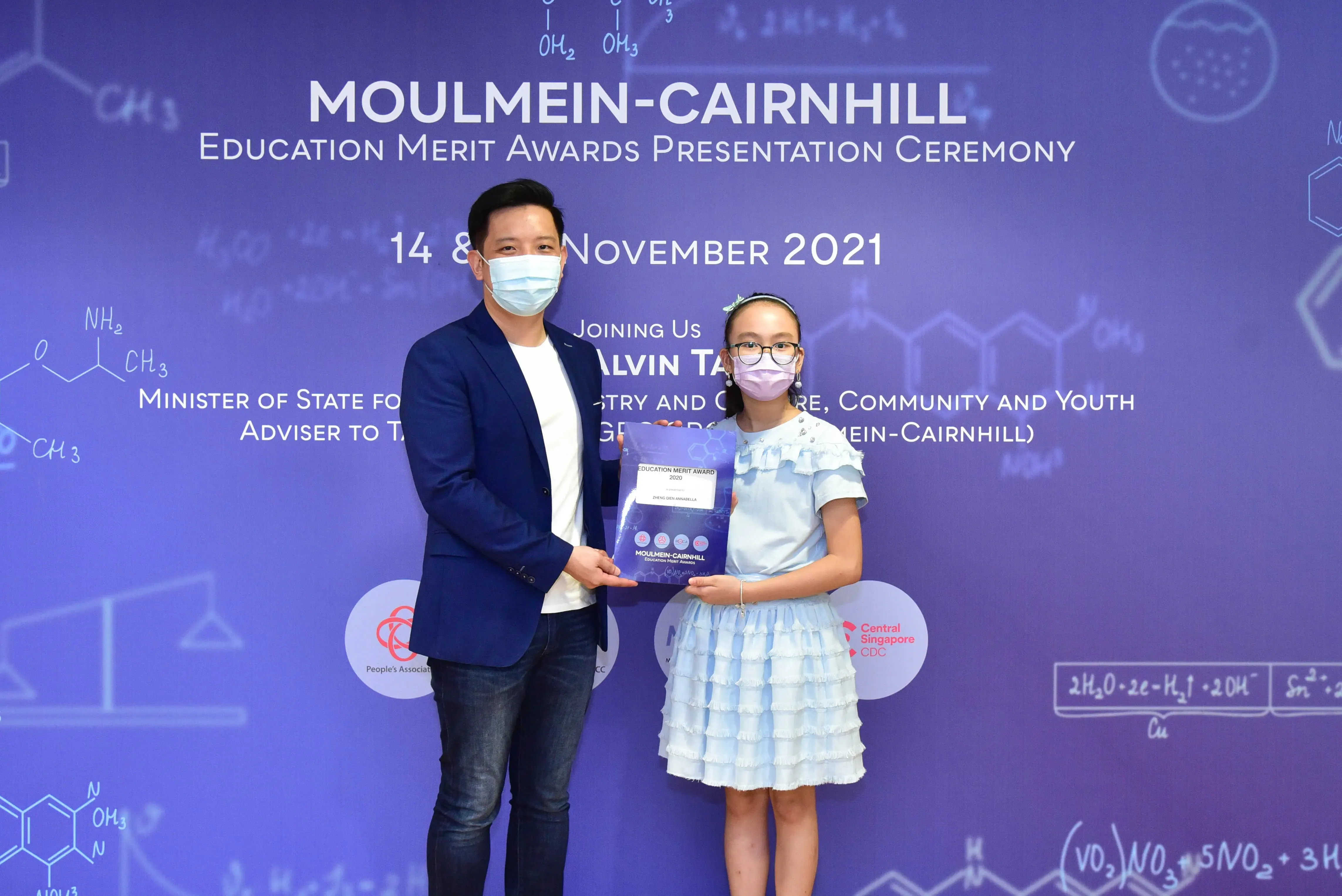 A man and a young girl in masks hold an award at the Moulmein-Cairnhill Education Merit Awards Ceremony, November 2021.