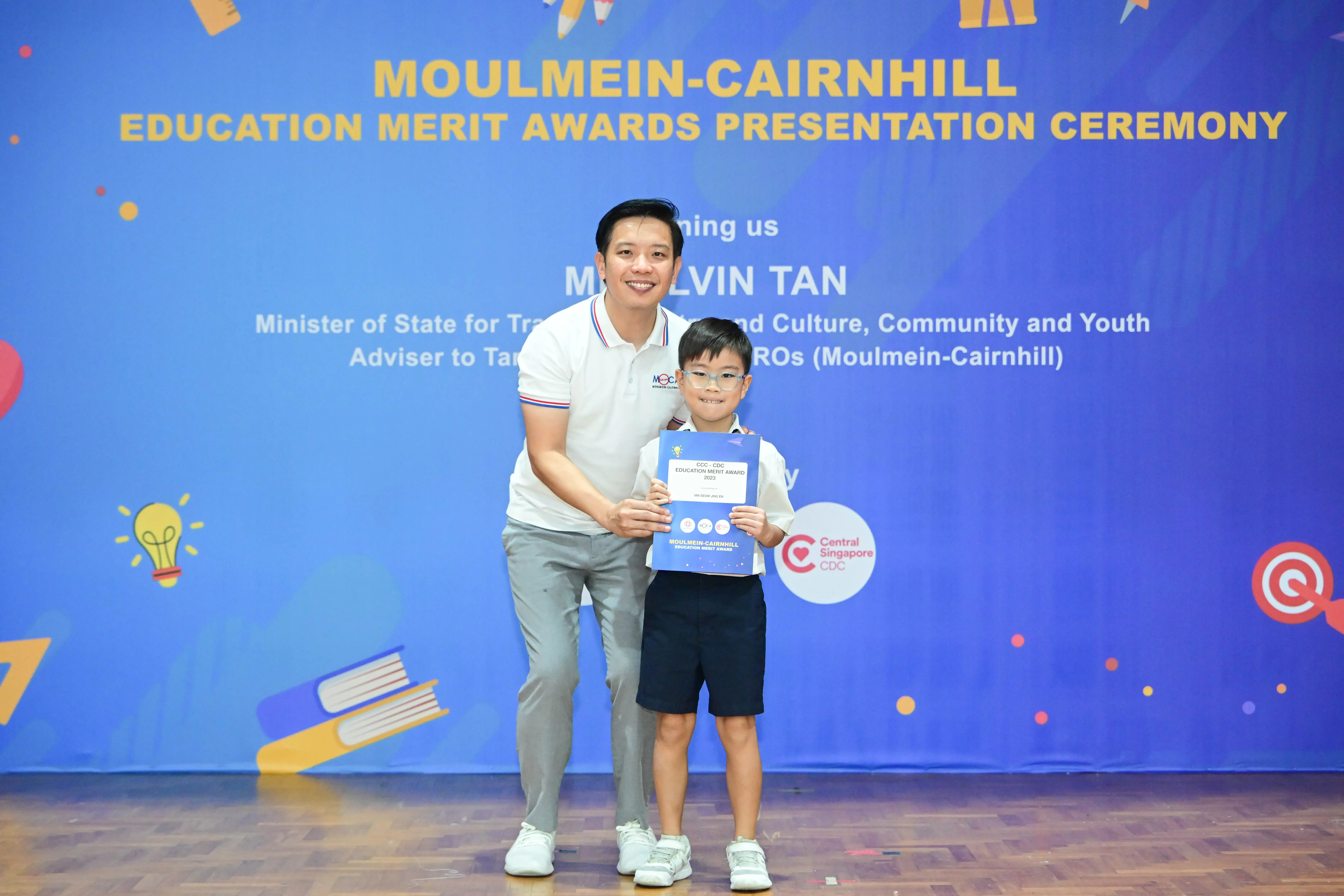 A man and a young boy holding a certificate pose before a Moulmein-Cairnhill Education Merit Awards banner.