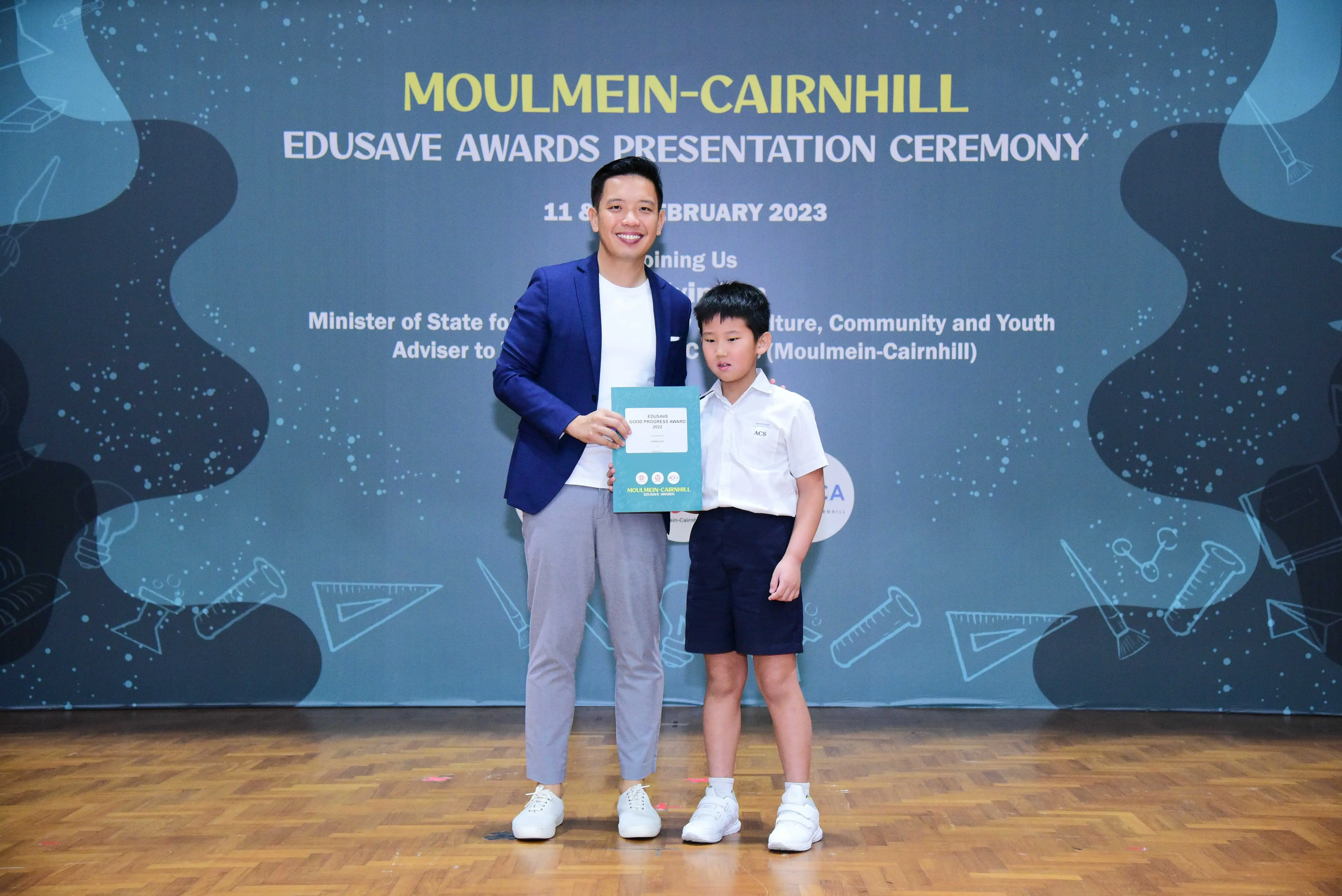 Adult in blue blazer and young boy in school uniform hold a certificate at Moulmein-Cairnhill Edusave Awards Ceremony 2023.
