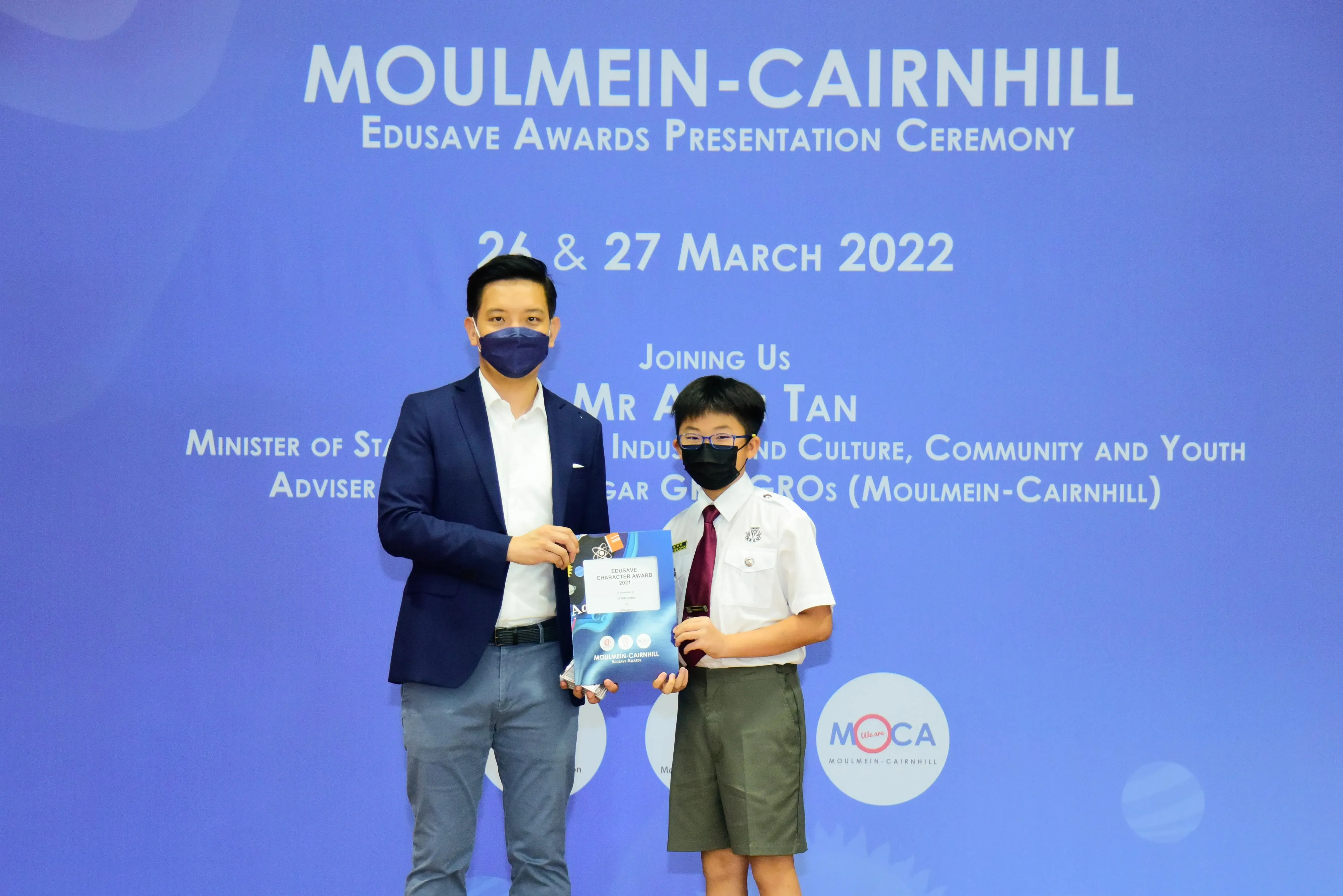 An adult and a masked student in school uniform hold an award at the Moulmein-Cairnhill Edusave Awards Ceremony 2022.