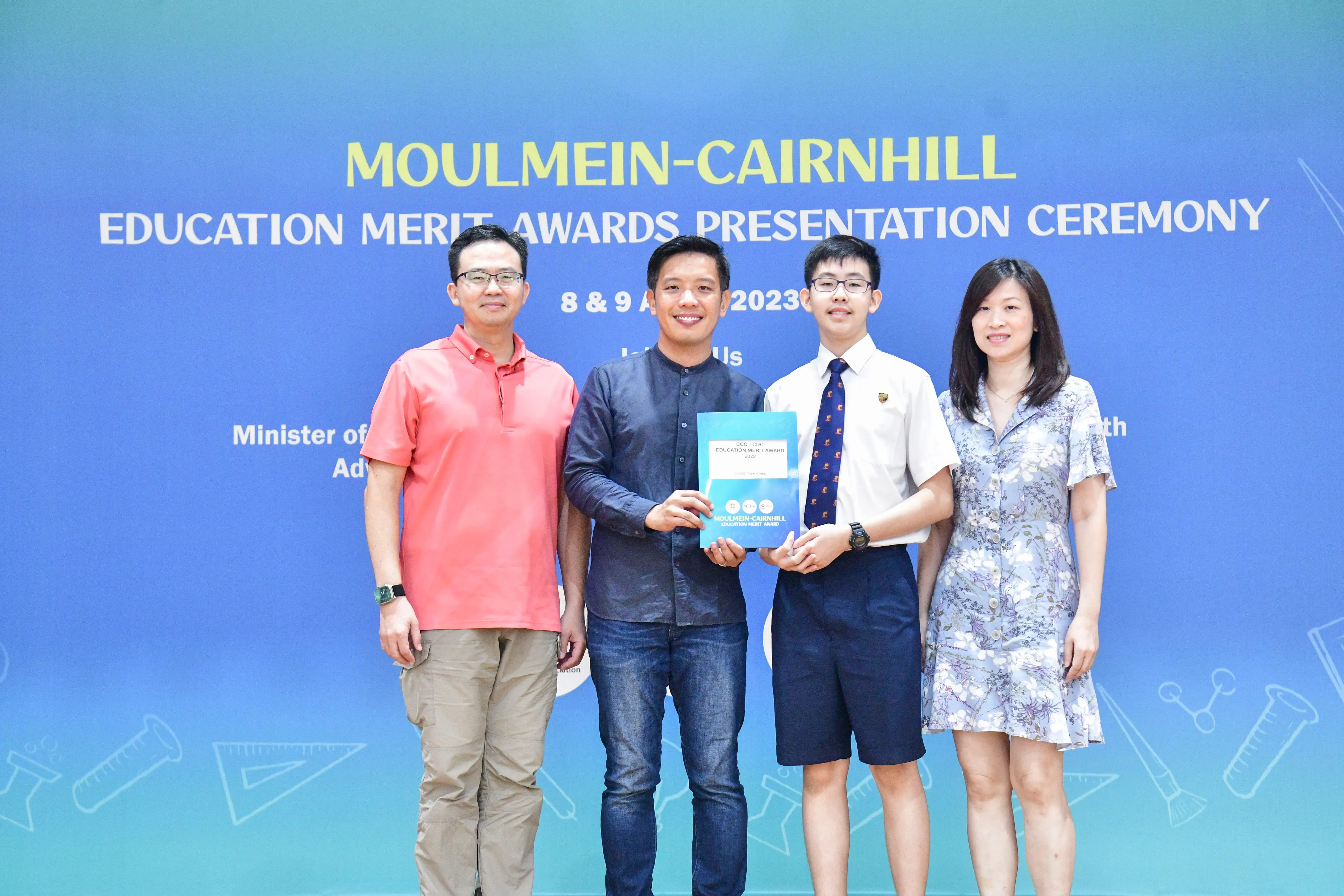 Four people pose at the Moulmein-Cairnhill Education Merit Awards Presentation Ceremony, holding a blue award folder.