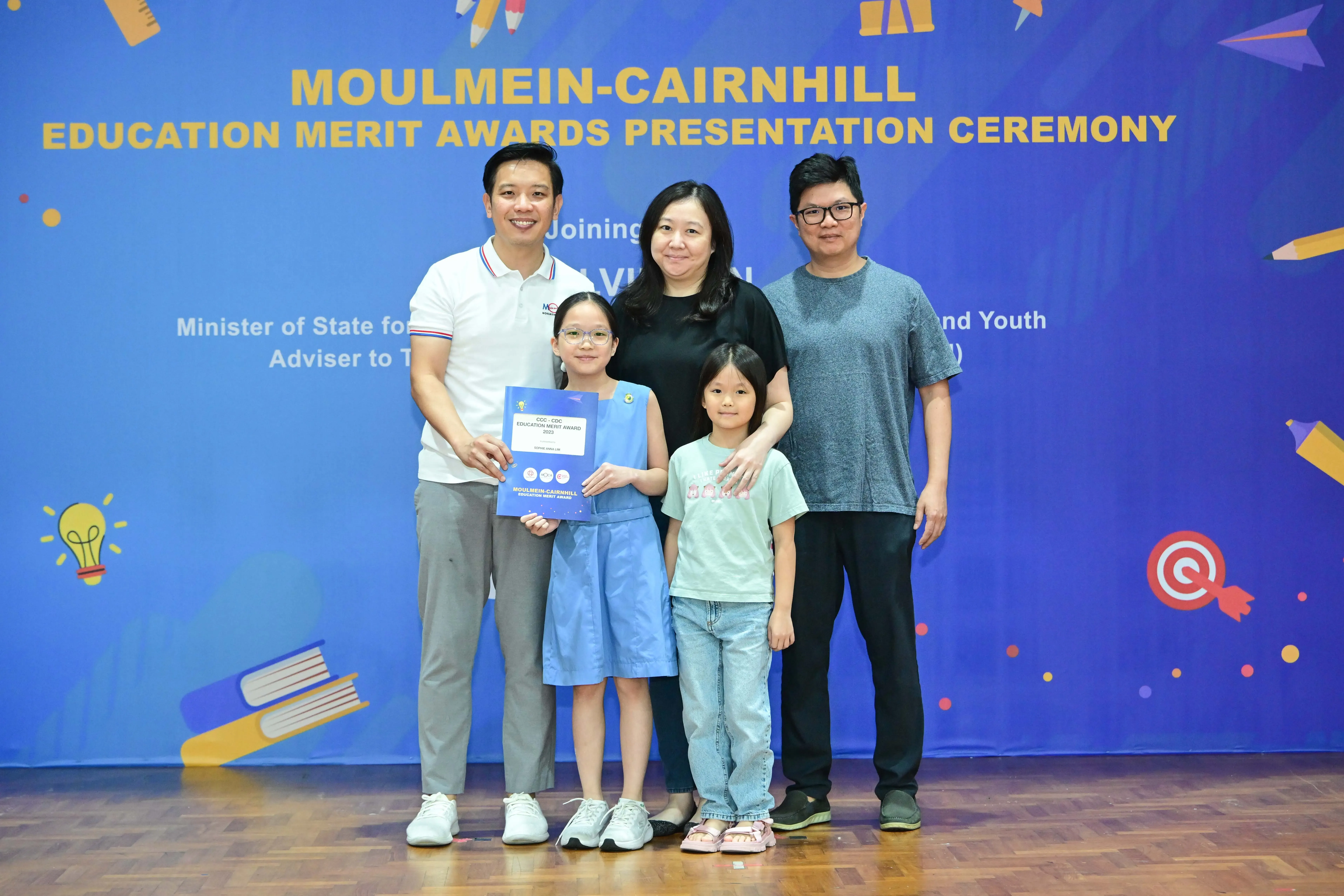Five people pose in front of a Moulmein-Cairnhill Education Merit Awards banner; a girl holds a certificate.