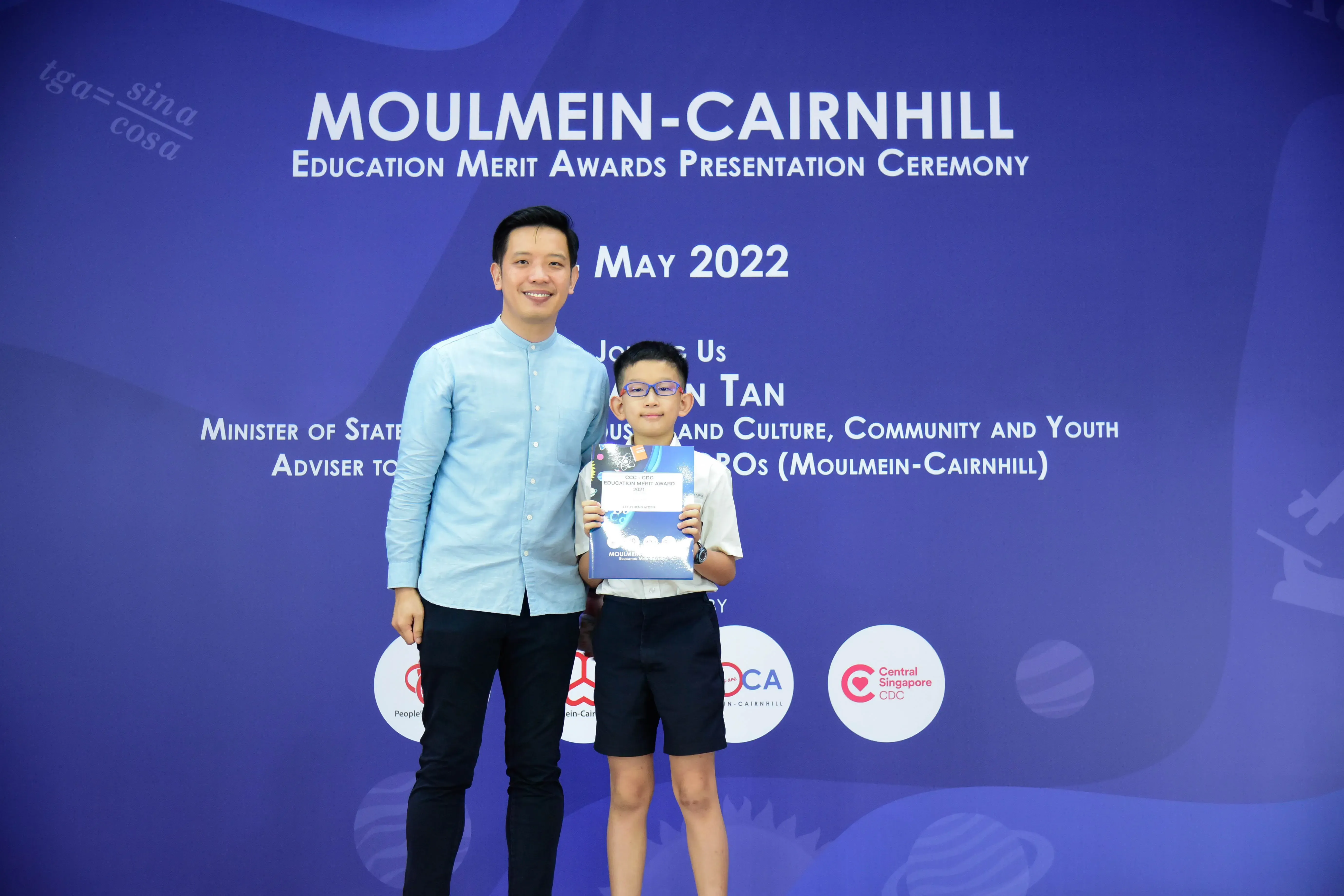 A man and a young boy holding an award certificate stand before a Moulmein-Cairnhill Education Merit Awards May 2022 backdrop.
