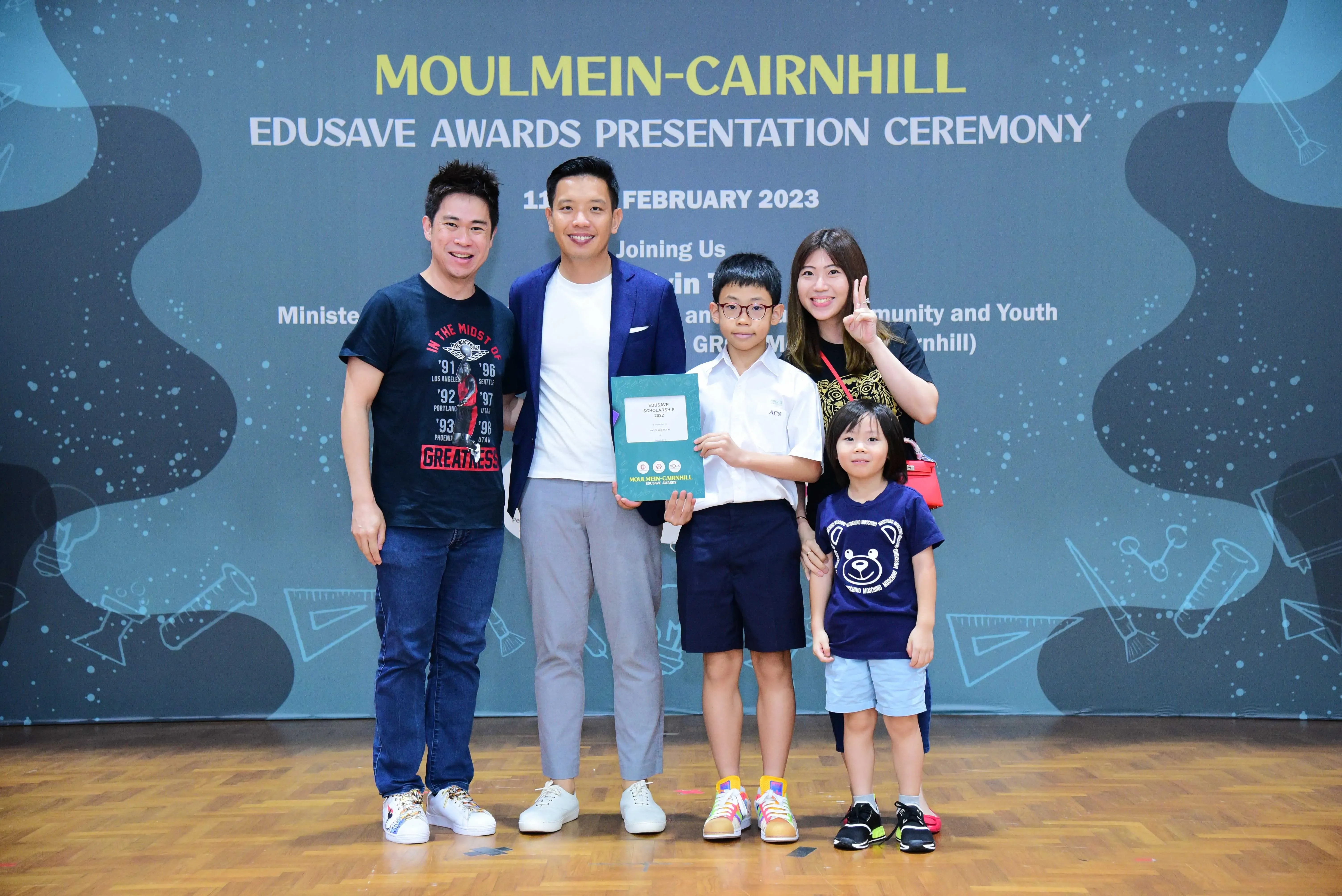 Five people pose before a Moulmein-Cairnhill Edusave Awards Presentation Ceremony banner dated February 2023.