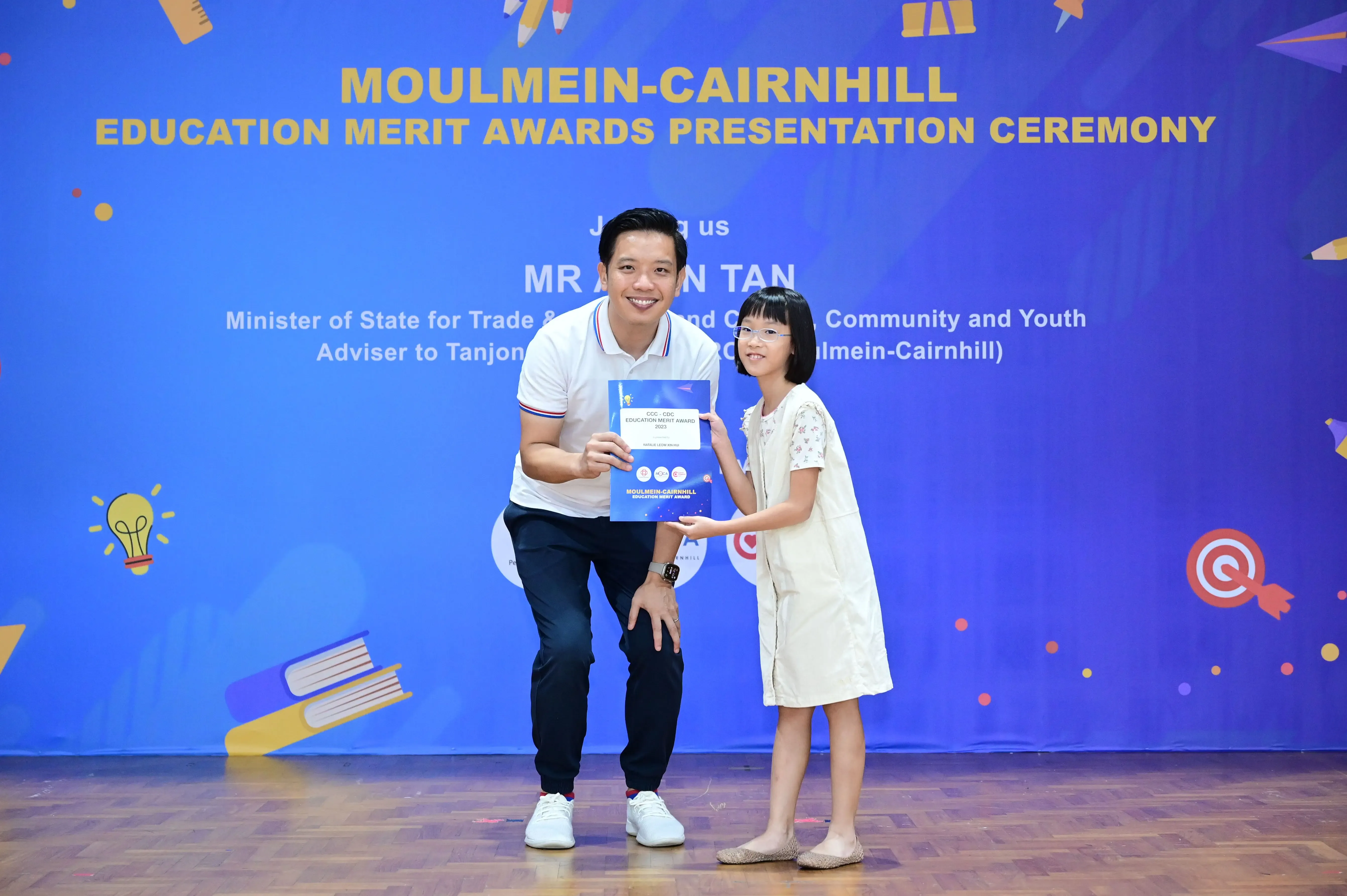 A man and a young girl hold a blue award certificate at the Moulmein-Cairnhill Education Merit Awards ceremony.