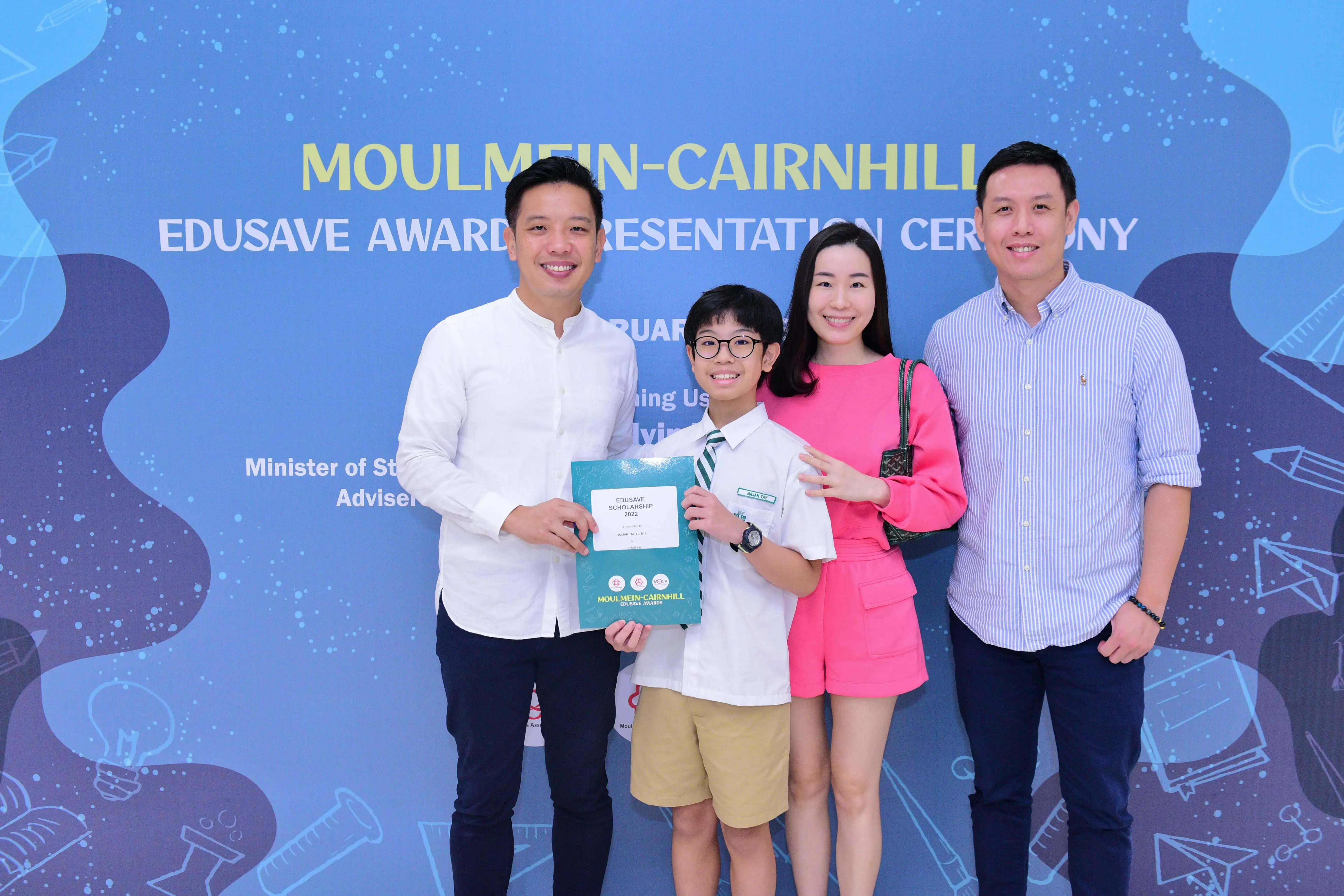 Four people smile before a Moulmein-Cairnhill Edusave Awards banner, with a boy holding a scholarship certificate.