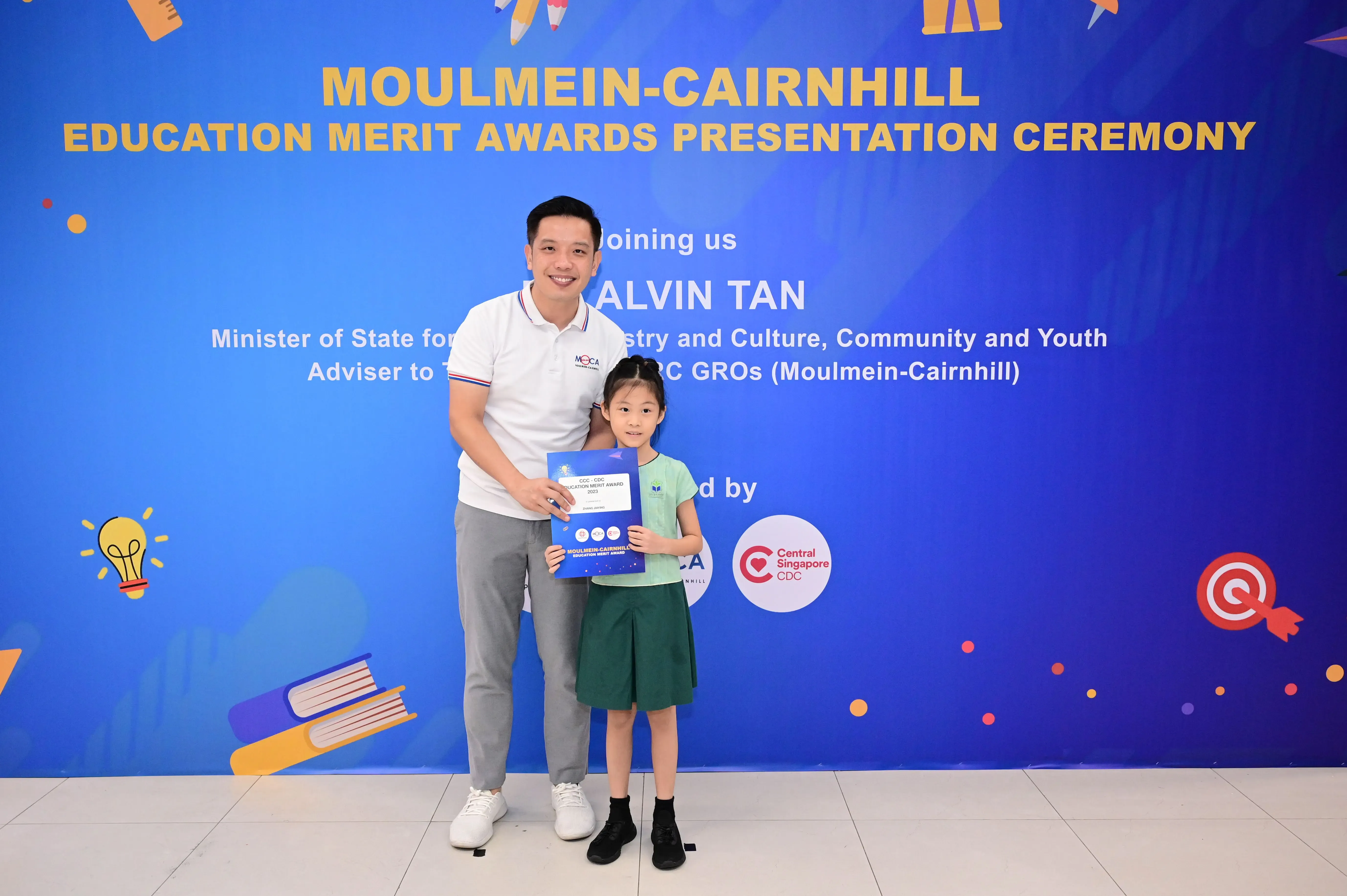 A man and a young girl hold an award certificate in front of a Moulmein-Cairnhill Education Merit Awards banner.