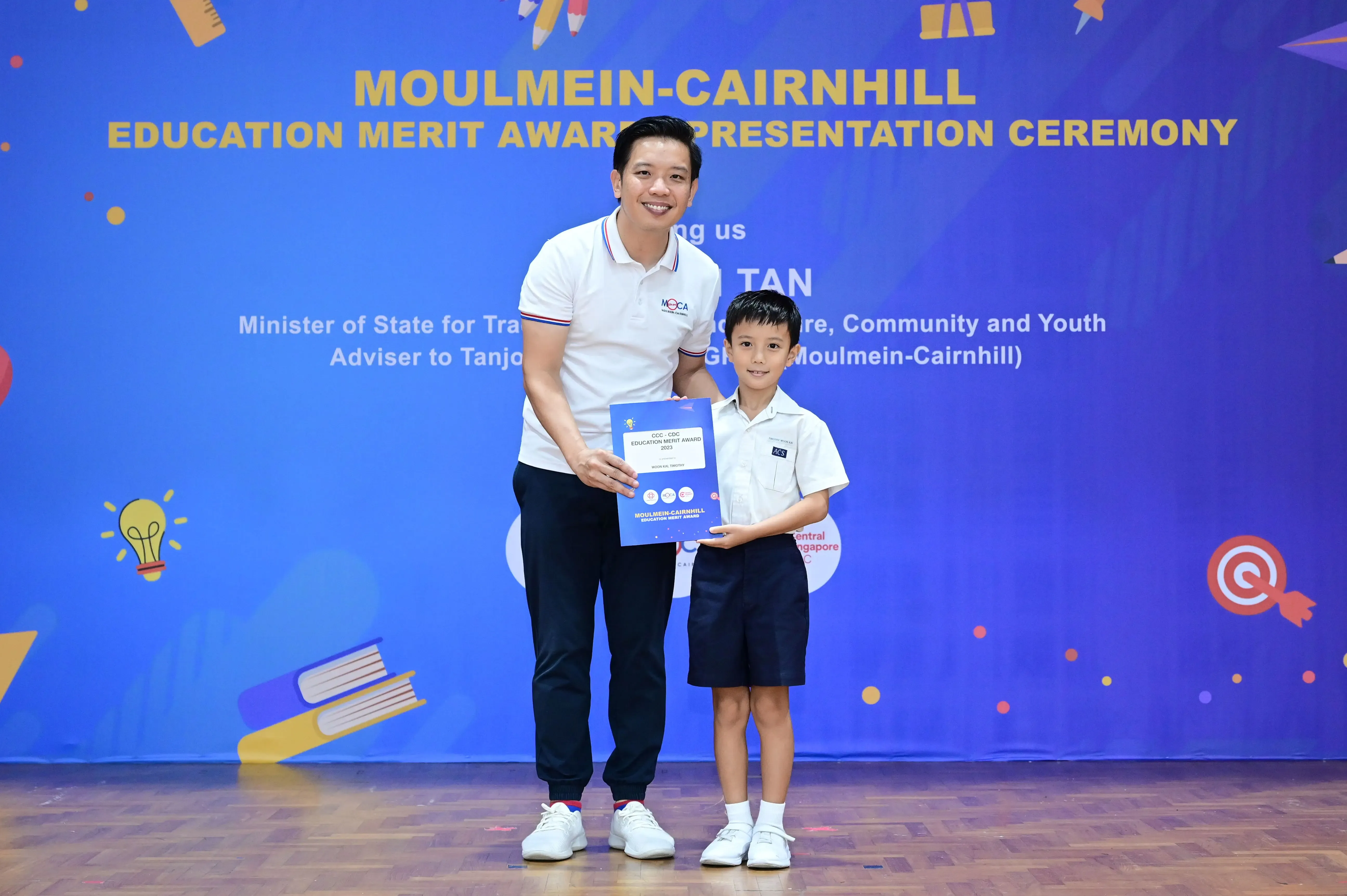 A man and a young boy hold a certificate together at the Moulmein-Cairnhill Education Merit Award Presentation Ceremony.