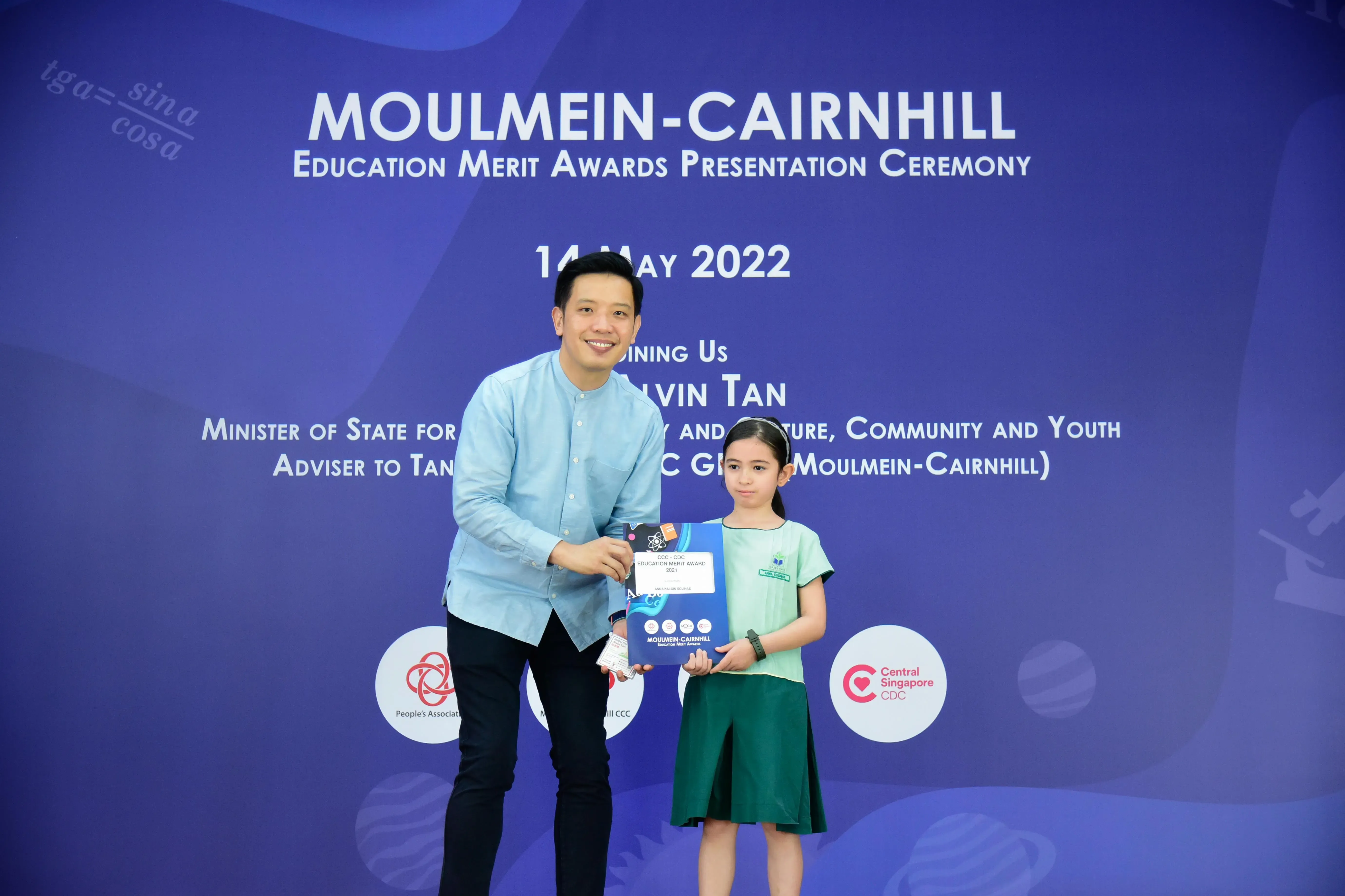 A man in blue shirt and a young girl in green uniform hold an award at the Moulmein-Cairnhill Education Merit Awards 2022.