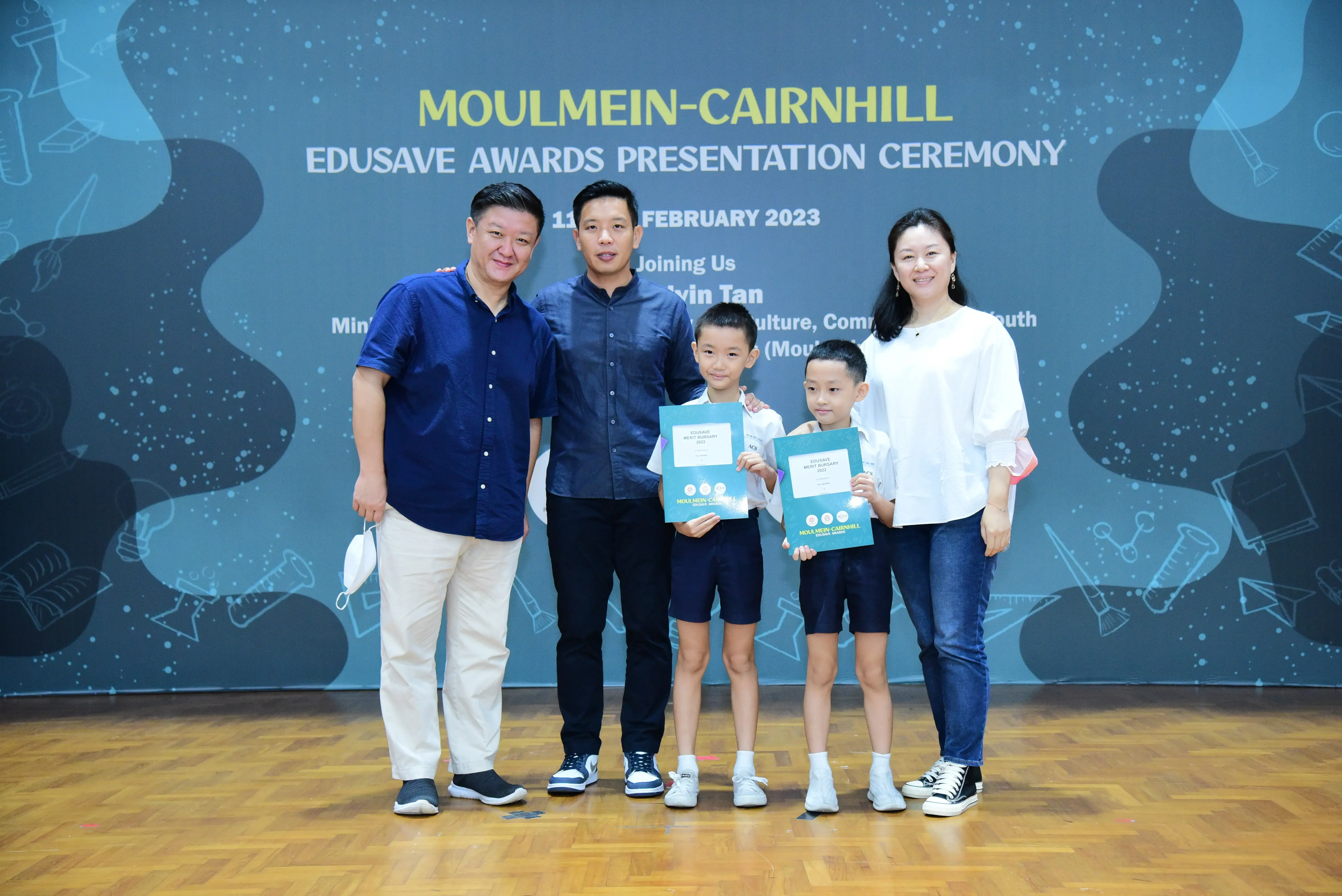 Five people pose at the Moulmein-Cairnhill Edusave Awards Presentation Ceremony, two children holding teal award folders.