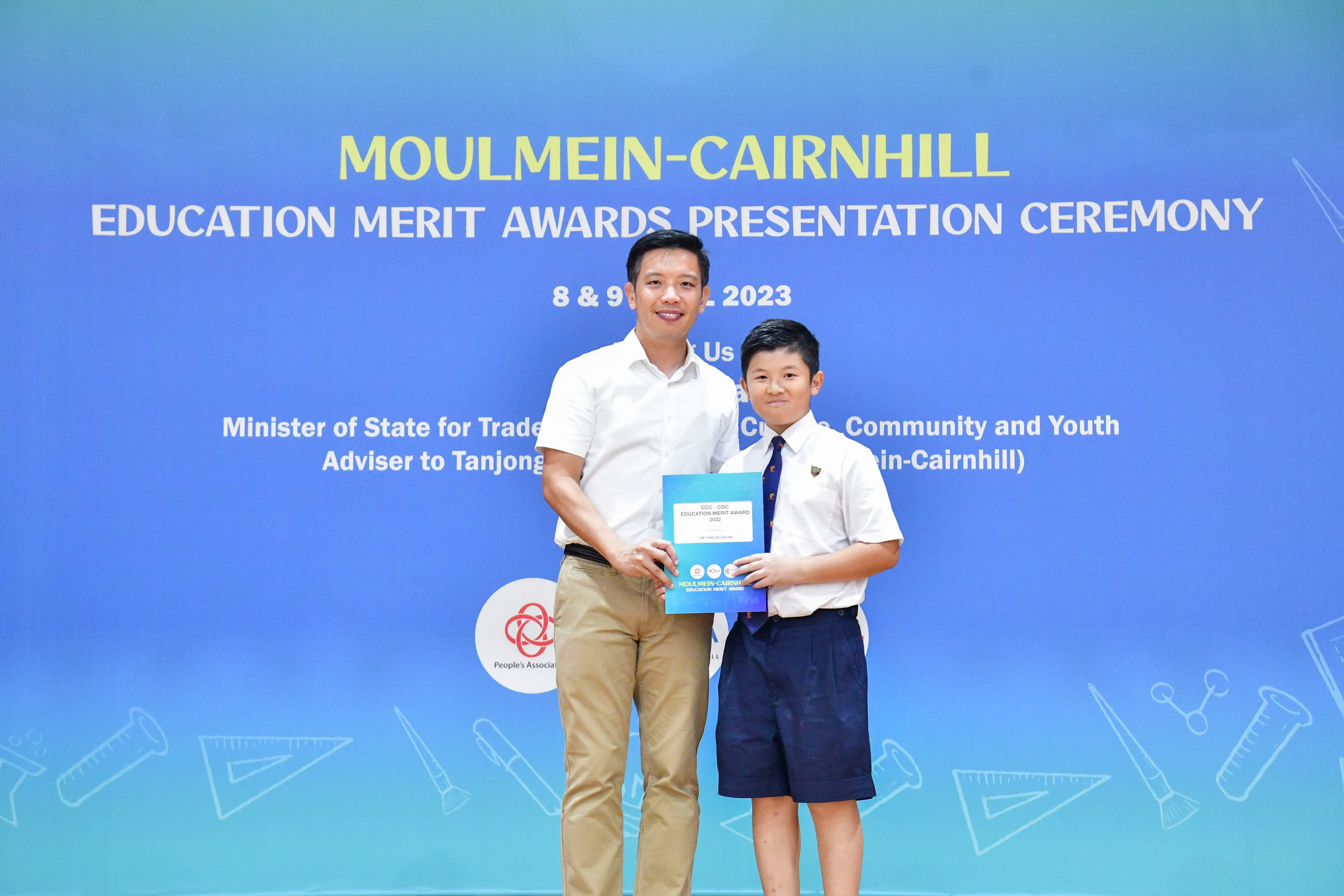 An adult man and a young boy in school uniform hold a certificate at the Moulmein-Cairnhill Education Merit Awards 2023.