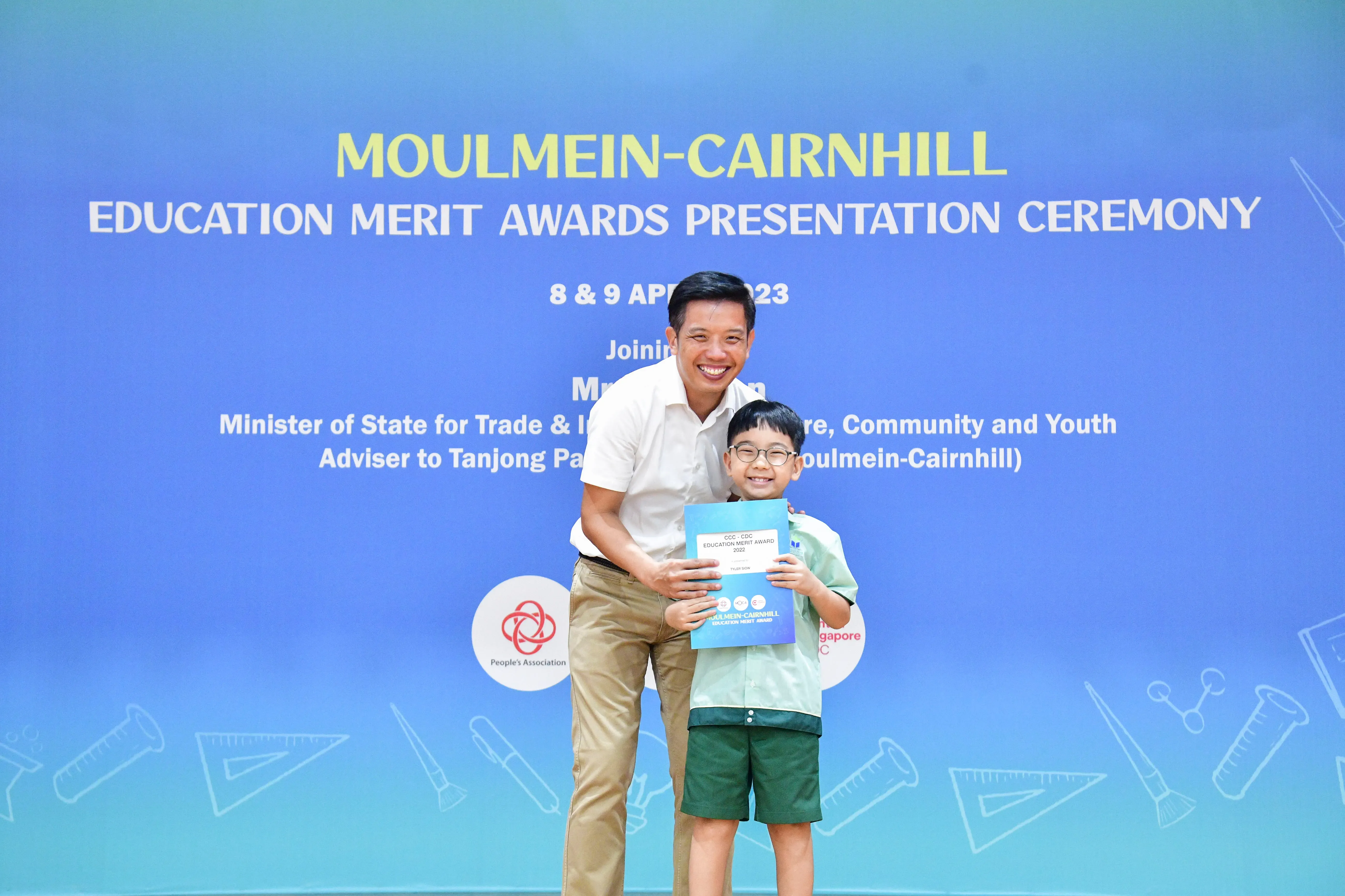 A man and a young boy holding a certificate stand before a Moulmein-Cairnhill Education Merit Awards banner.