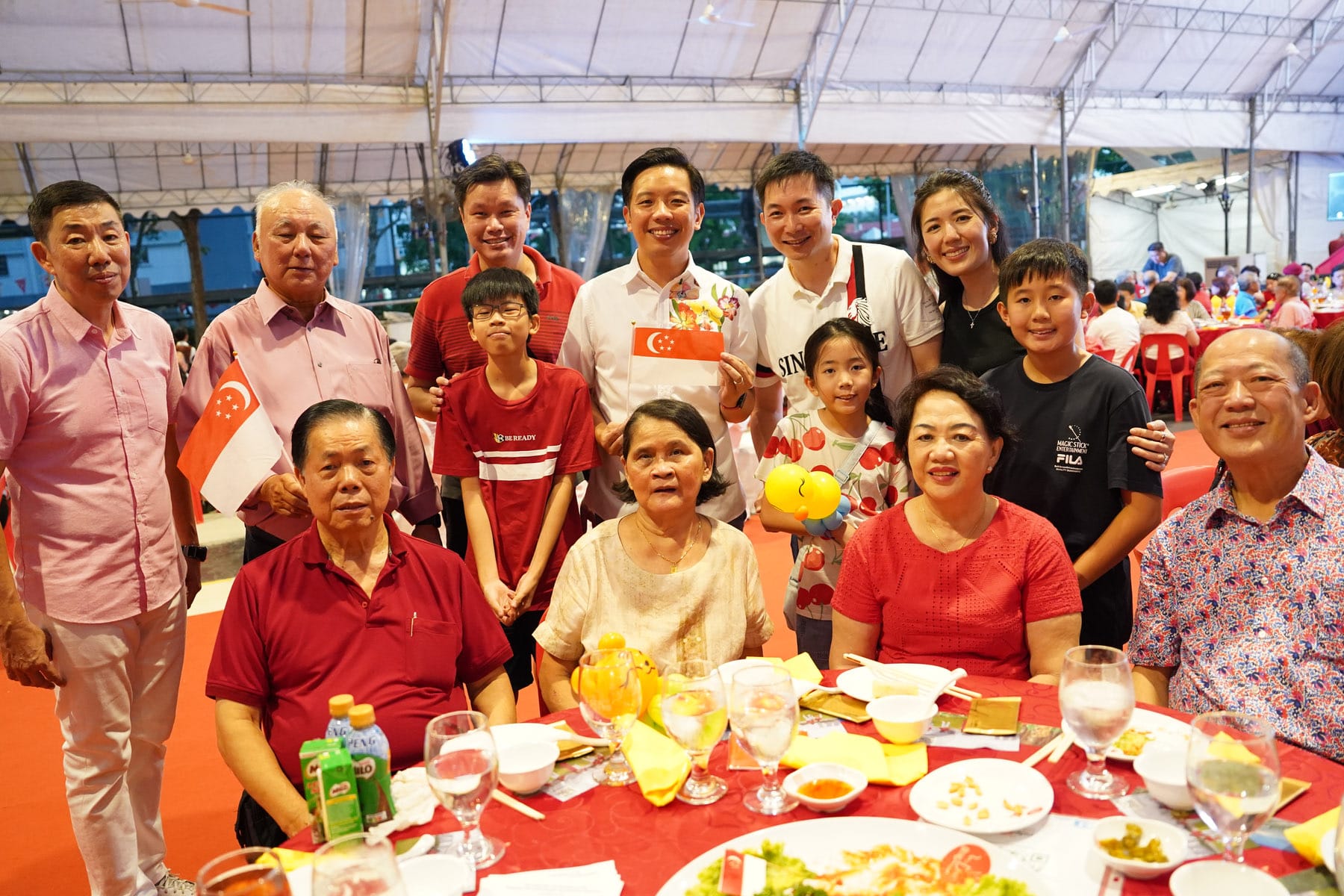 A large group of people posing together at a dinner event, some holding small Singapore flags, seated and standing around a red-clothed table.