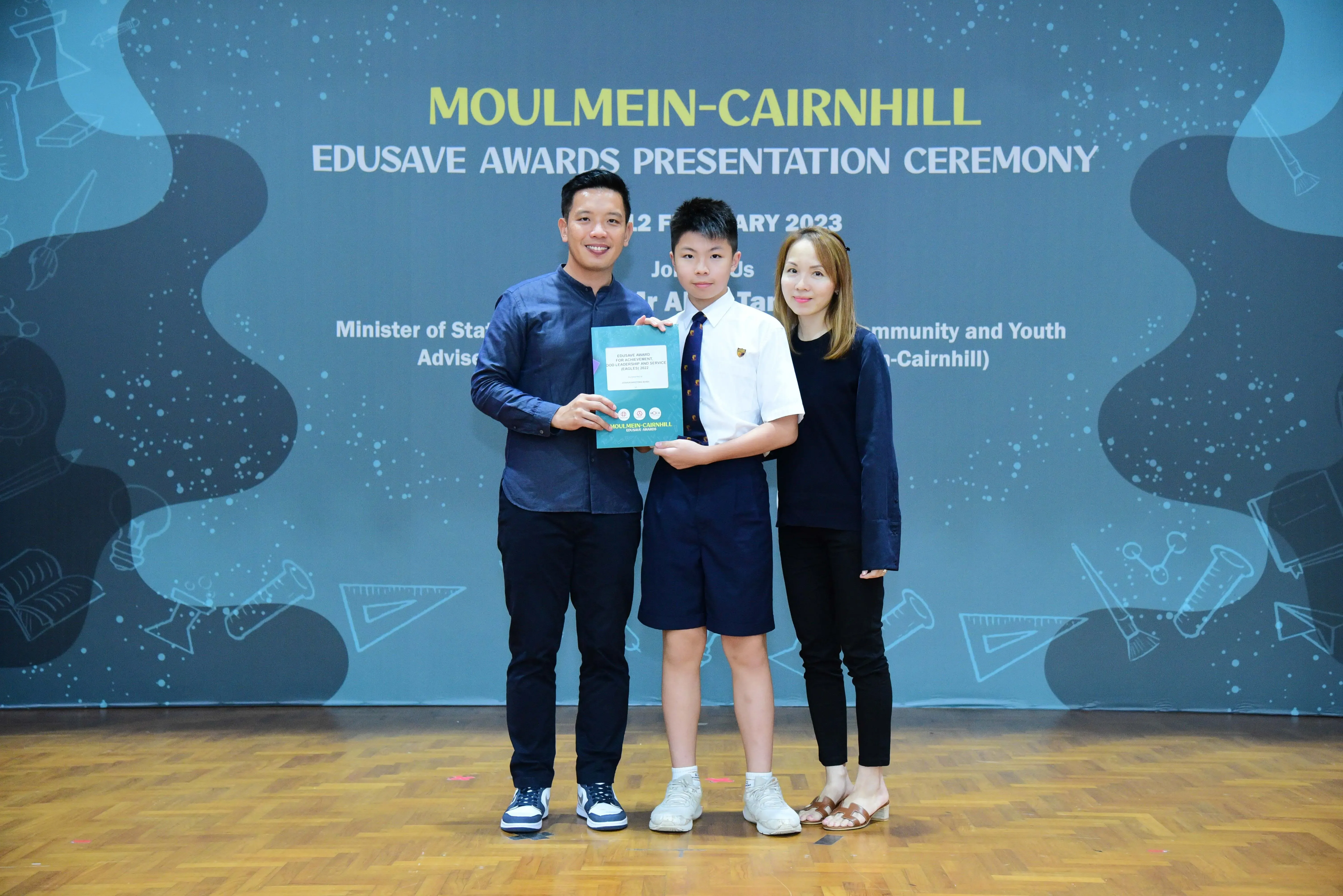 A boy in school uniform holds an award certificate flanked by two adults at the Moulmein-Cairnhill Edusave Awards 2023.