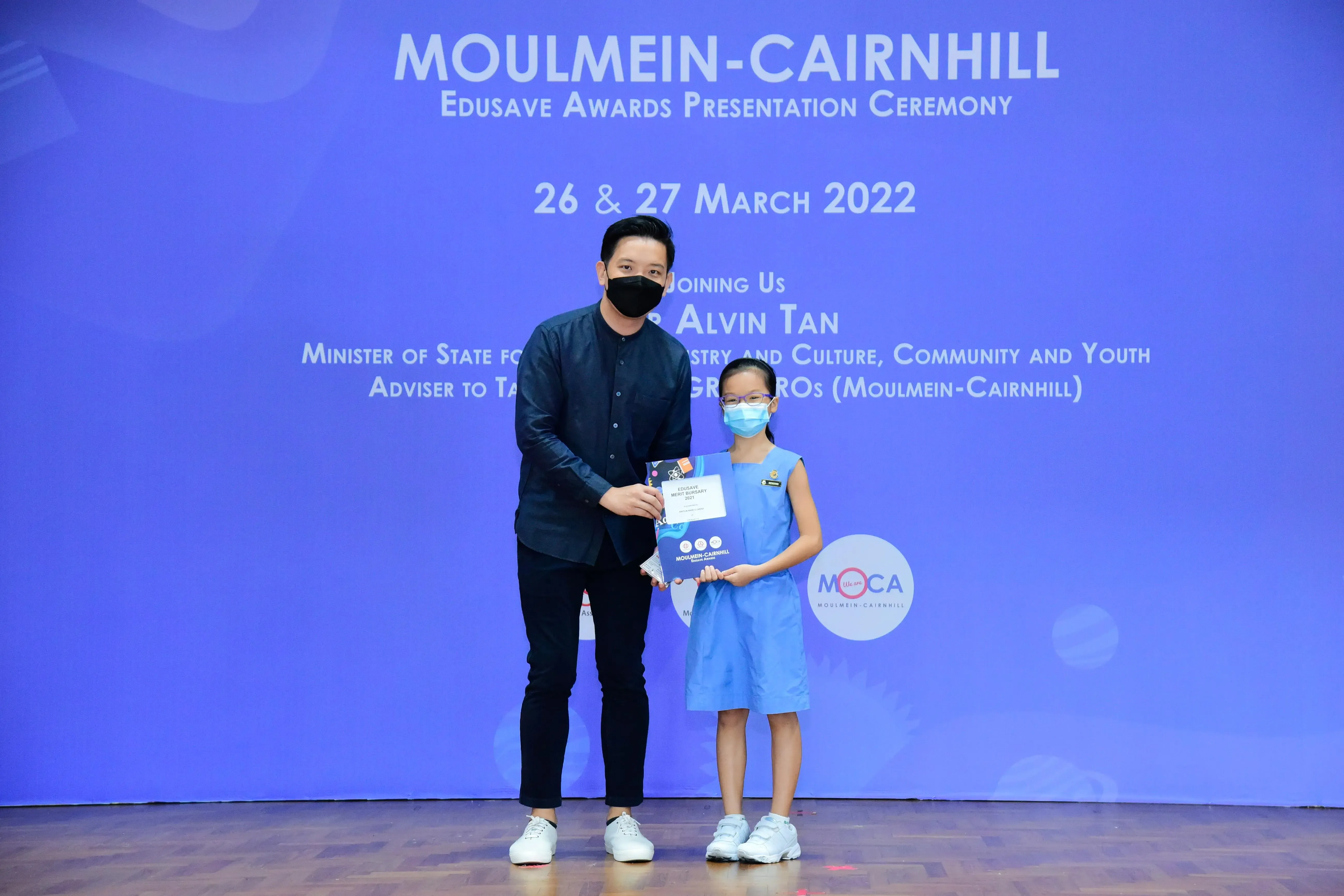 A masked man and young girl in blue dress hold a certificate at the Moulmein-Cairnhill Edusave Awards Ceremony 2022.