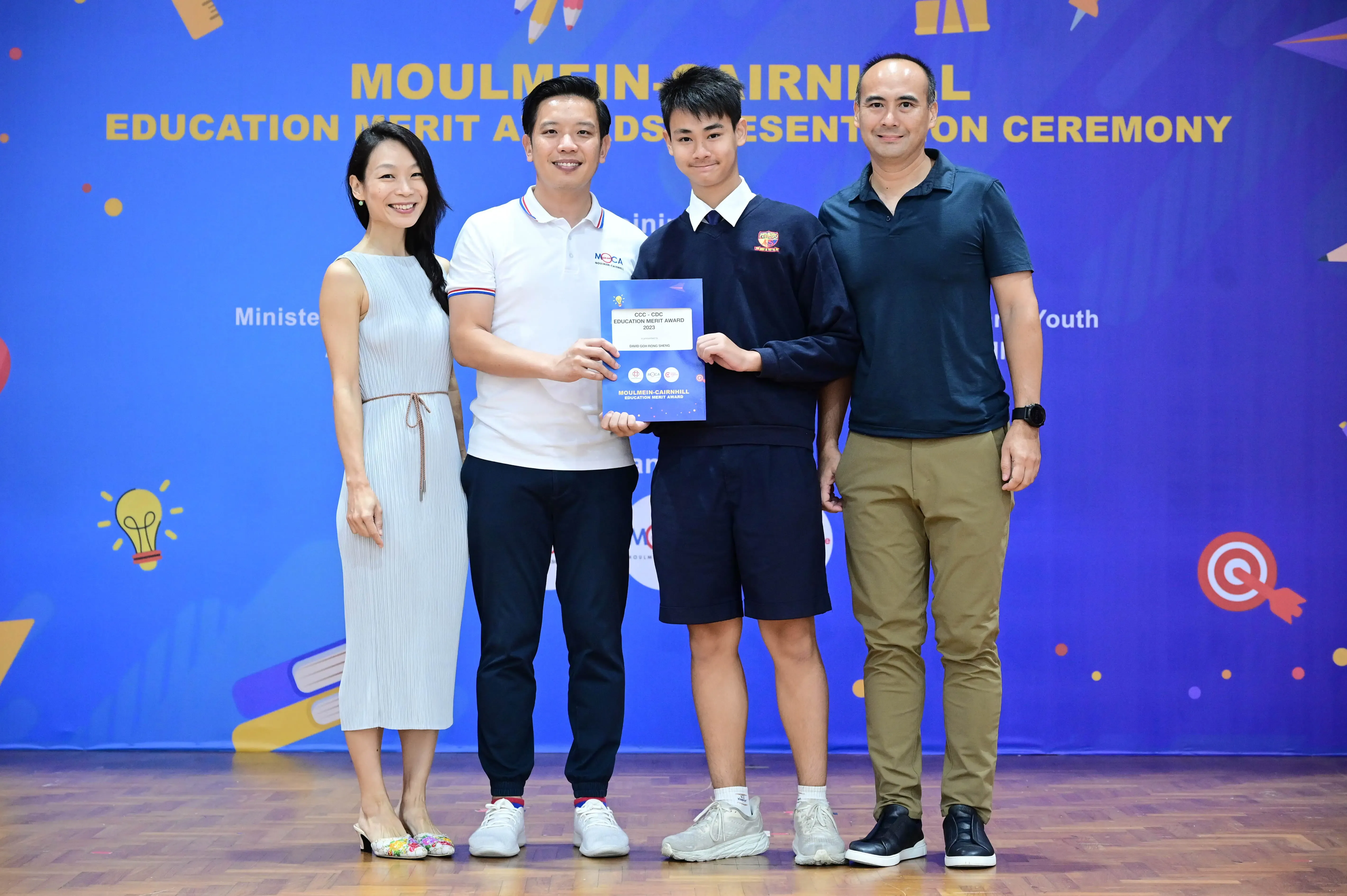 Four people pose together at the Moulmein-Cairnhill Education Merit Awards Presentation Ceremony, holding an award certificate.