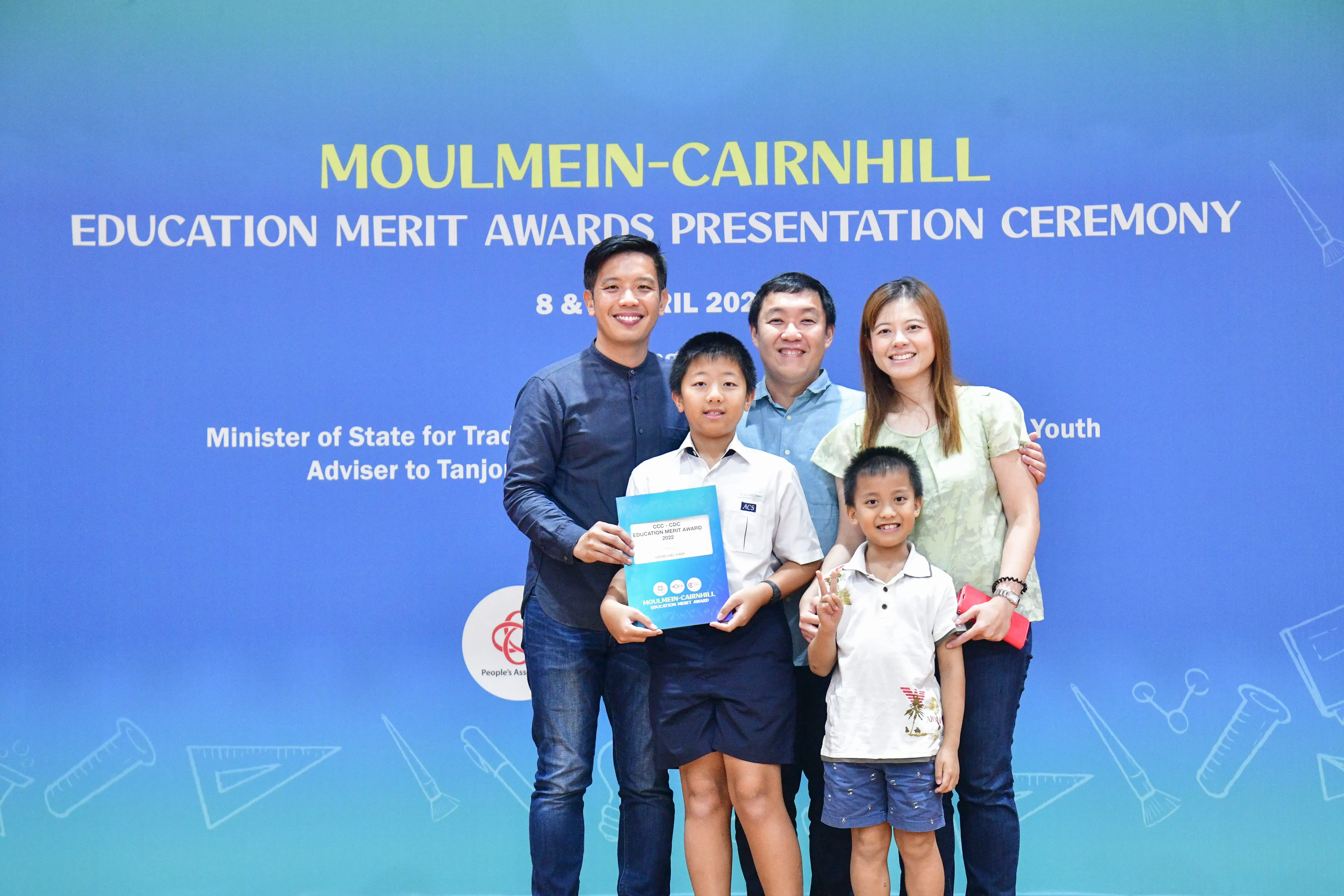 Five people pose before a Moulmein-Cairnhill Education Merit Awards Presentation Ceremony banner, a boy holding an award folder.