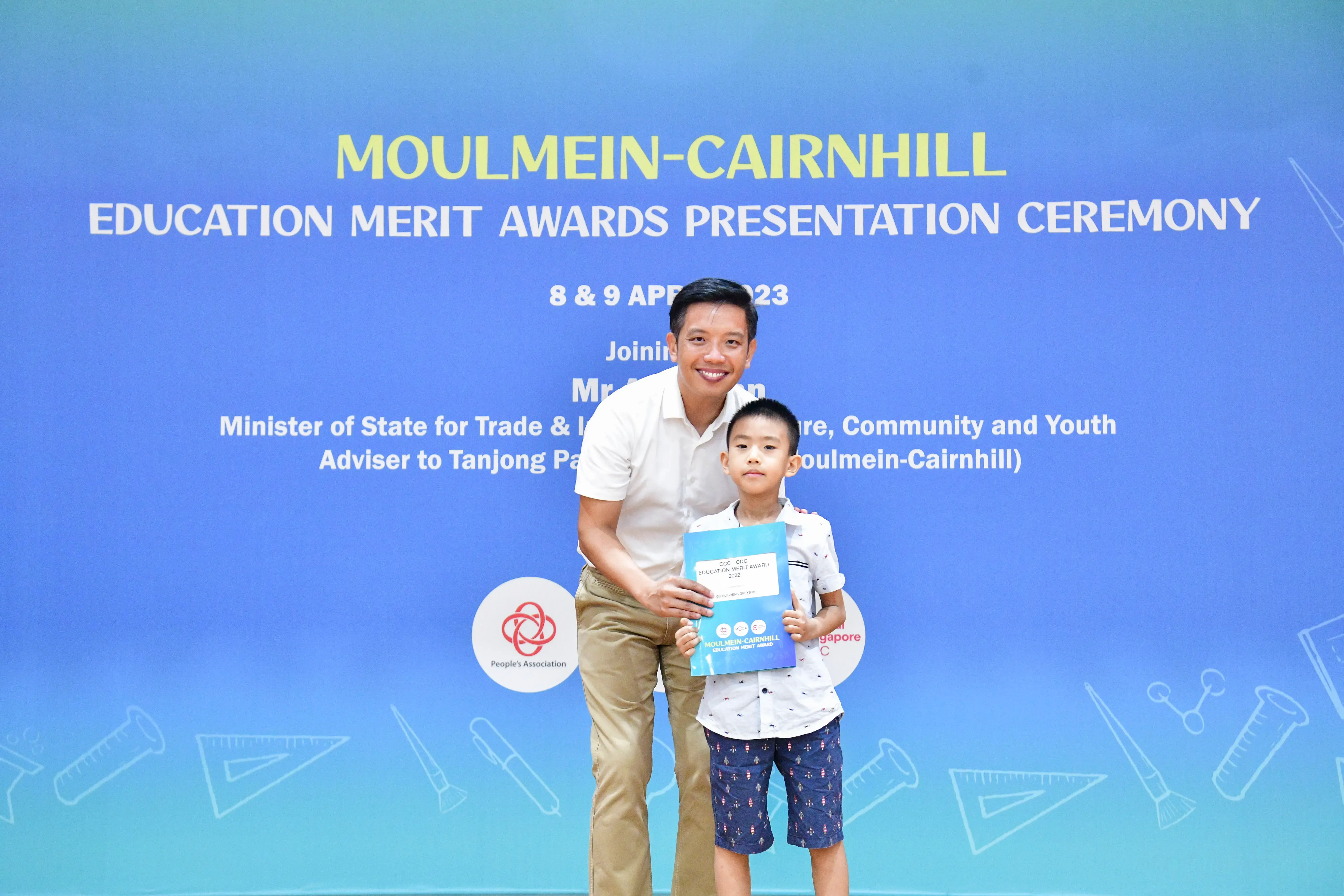 A man and a young boy holding an award certificate stand before a Moulmein-Cairnhill Education Merit Awards banner.