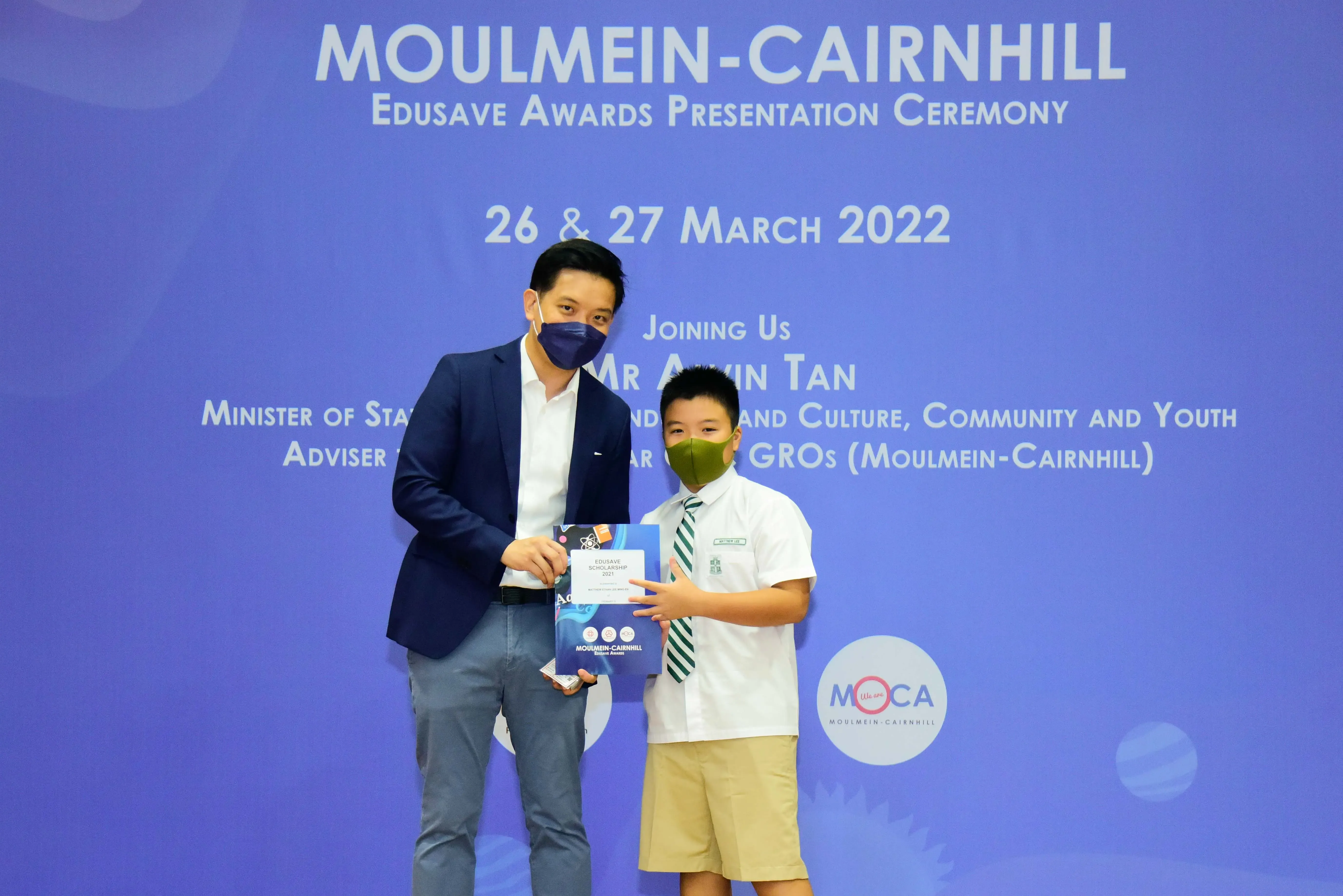 An adult and a young student wearing masks hold an Edusave Scholarship at the Moulmein-Cairnhill Awards Ceremony 2022.