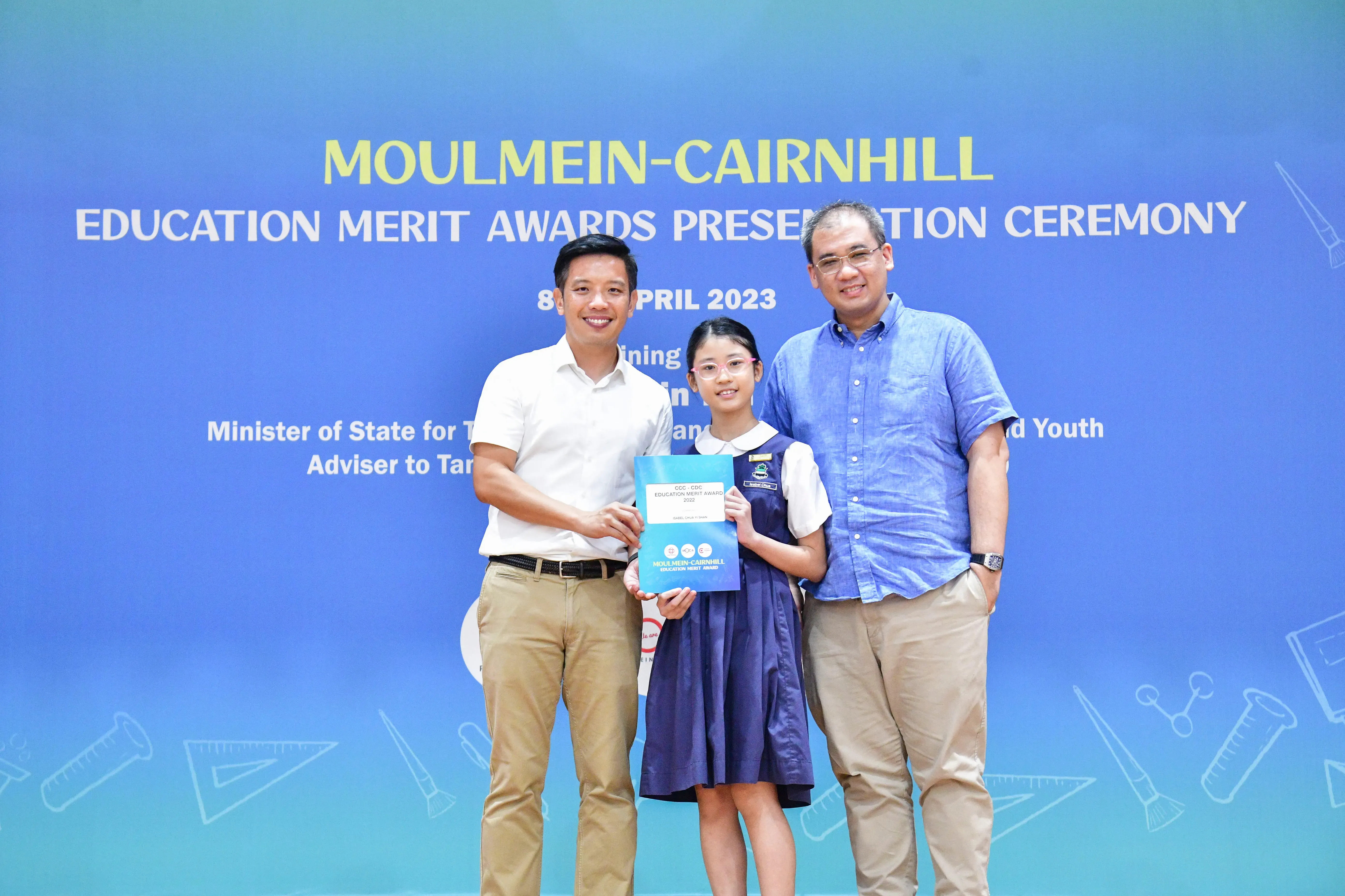 A girl holding an award certificate stands between two men at the Moulmein-Cairnhill Education Merit Awards 2023.