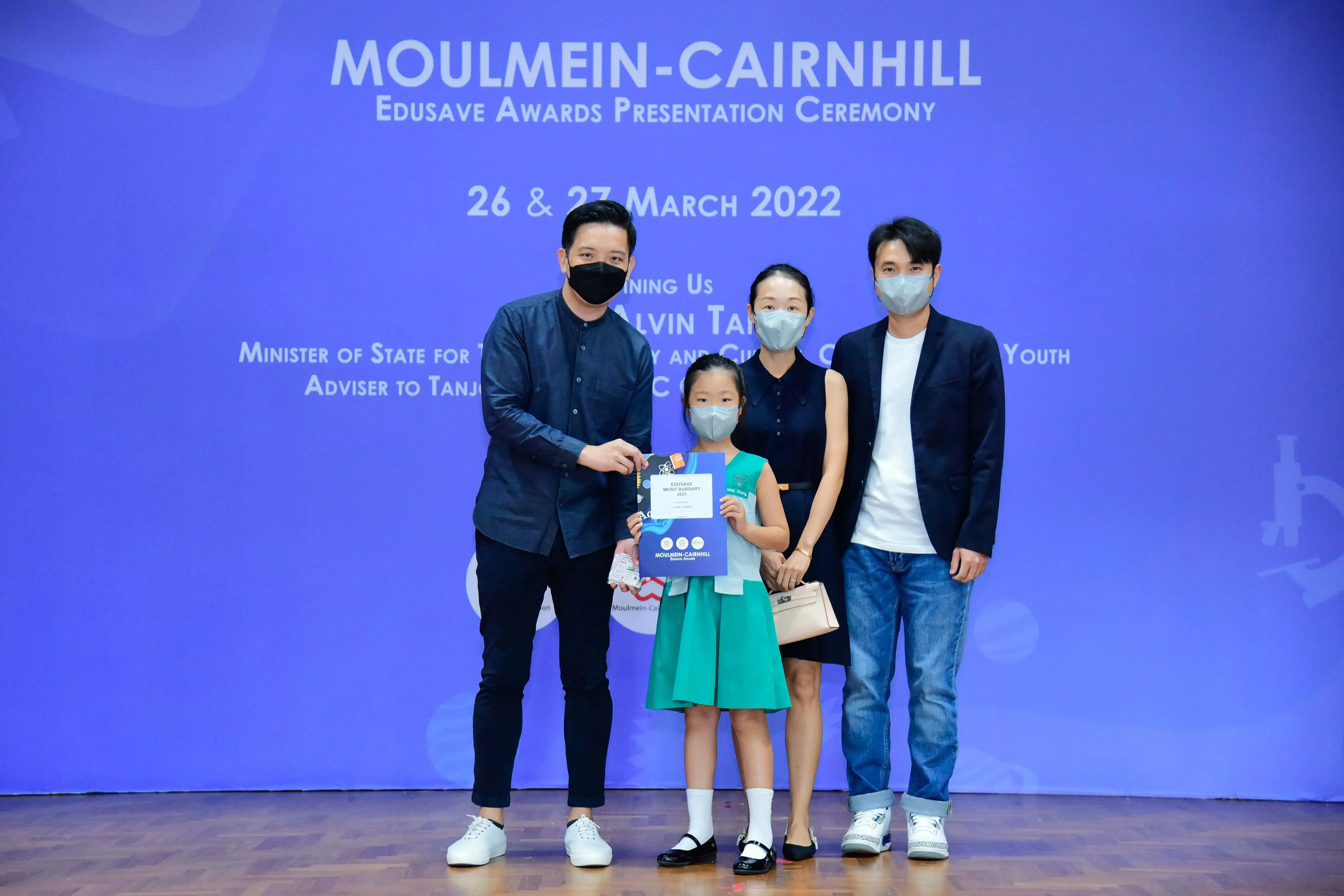 Four masked people pose at the Moulmein-Cairnhill Edusave Awards Ceremony on 26-27 March 2022, with a child holding a certificate.