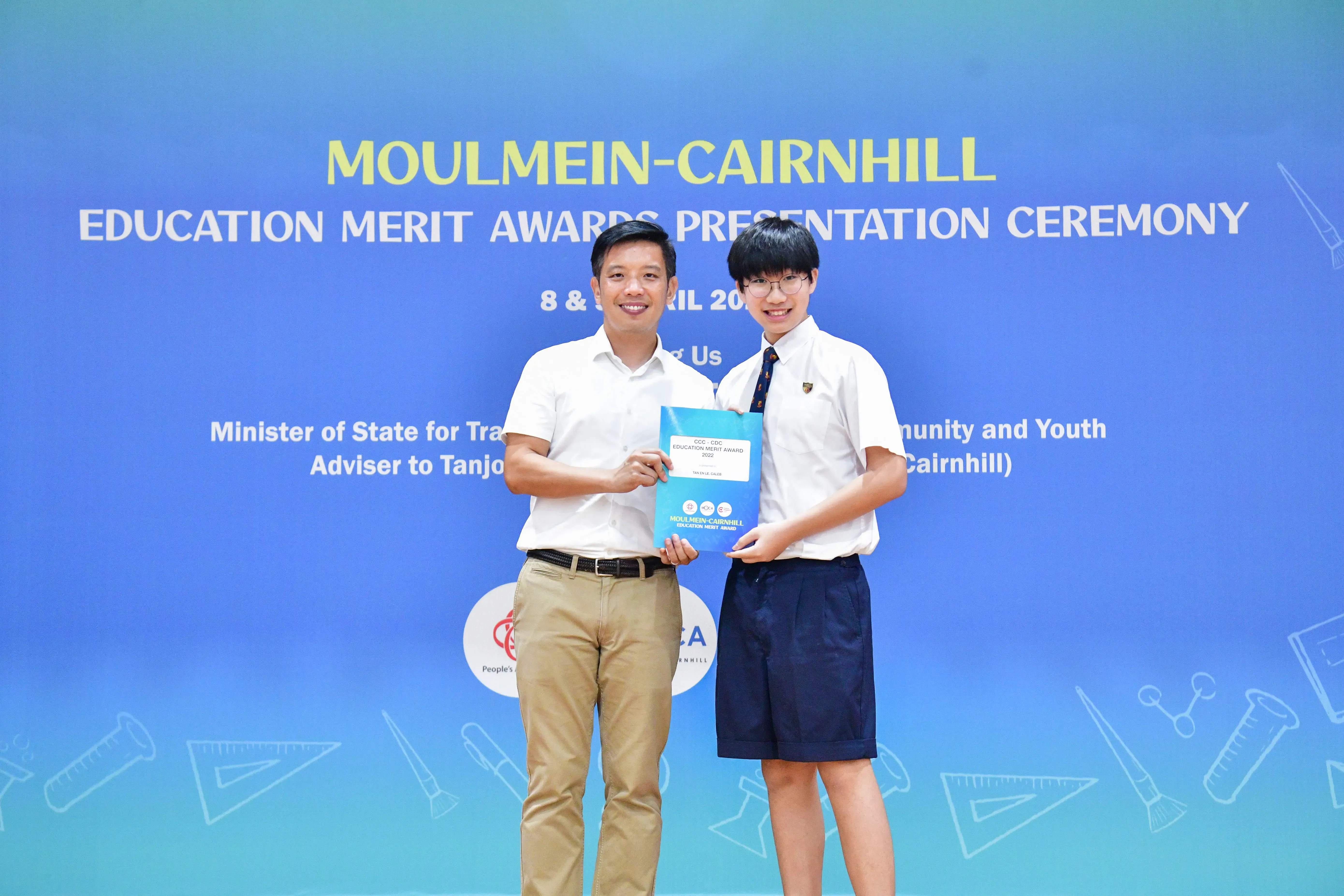 Two people hold a certificate together in front of a Moulmein-Cairnhill Education Merit Awards Presentation Ceremony banner.