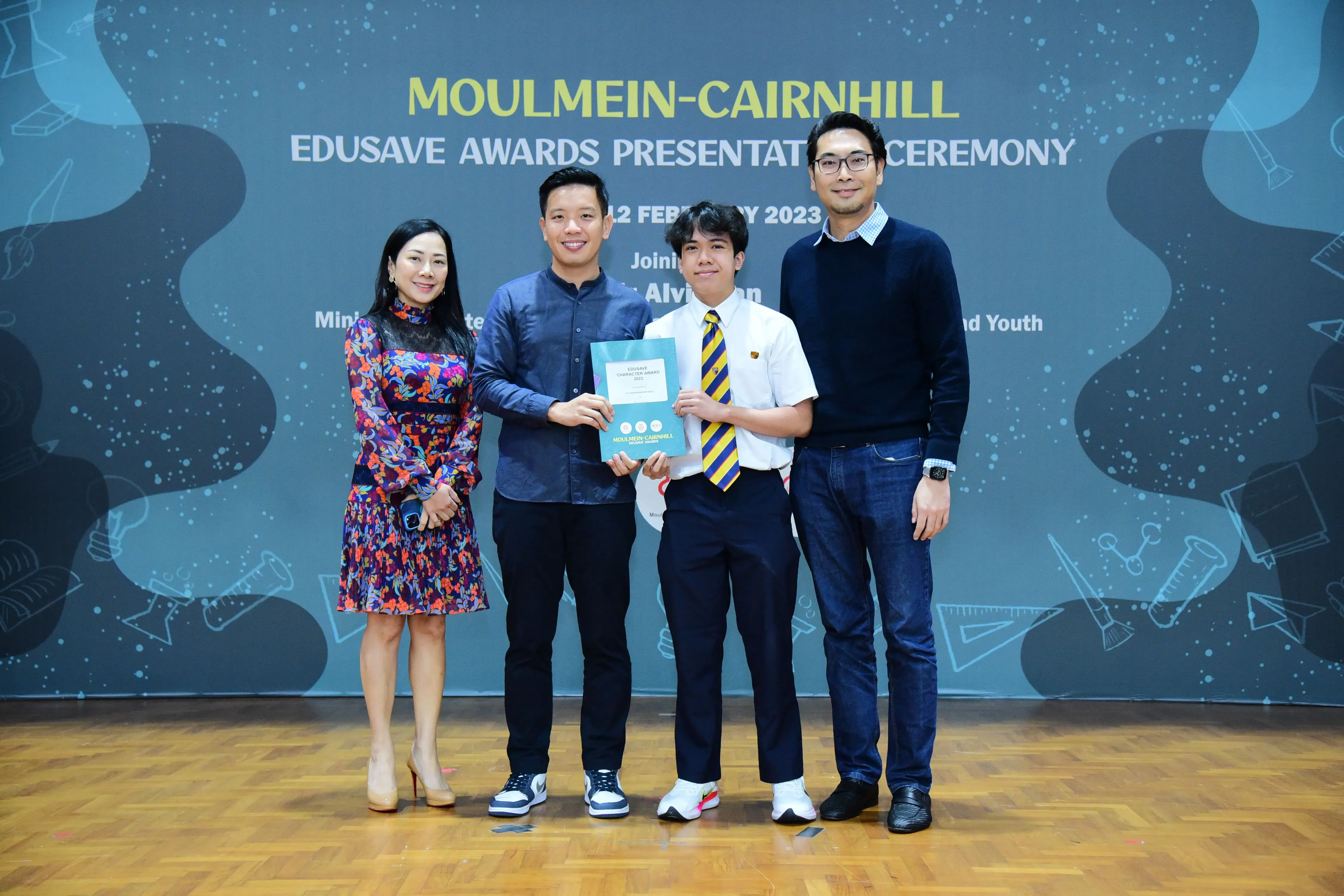 Four people pose on a stage at the Moulmein-Cairnhill Edusave Awards Ceremony 2023, holding a certificate.
