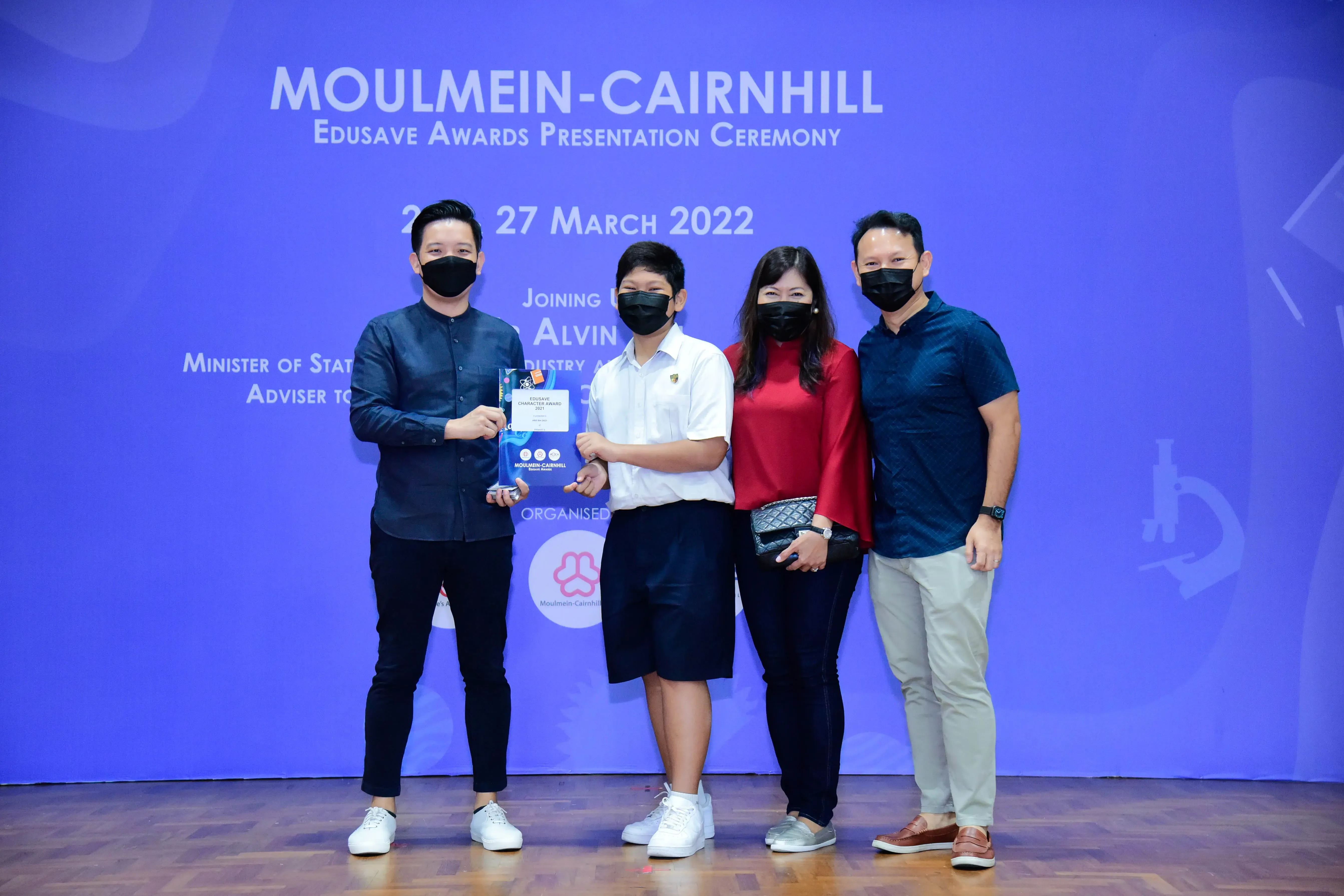 Four masked people pose at the Moulmein-Cairnhill Edusave Awards Ceremony on 27 March 2022, holding a trophy.