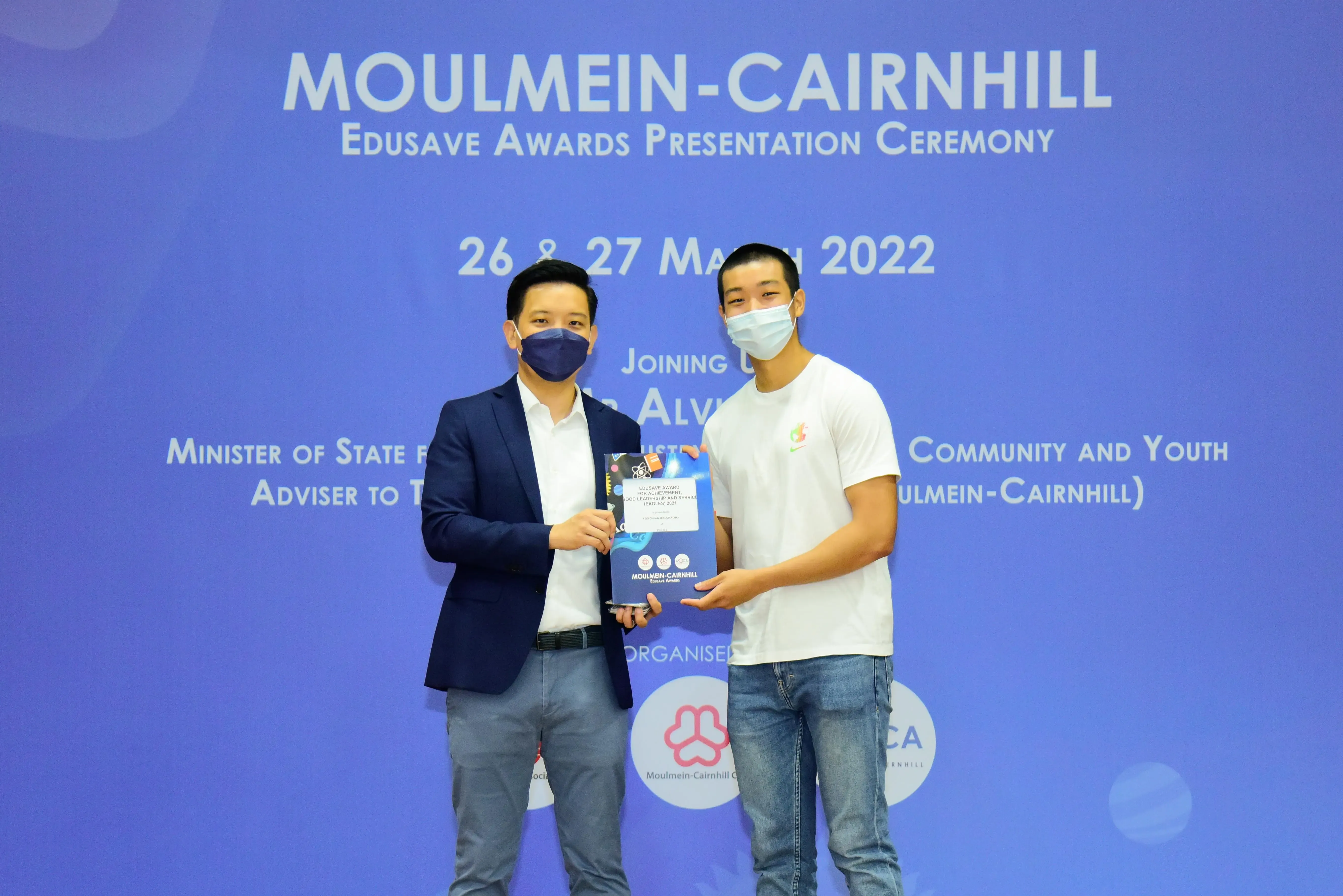 Two masked men hold an award at the Moulmein-Cairnhill Edusave Awards Presentation Ceremony, March 2022.