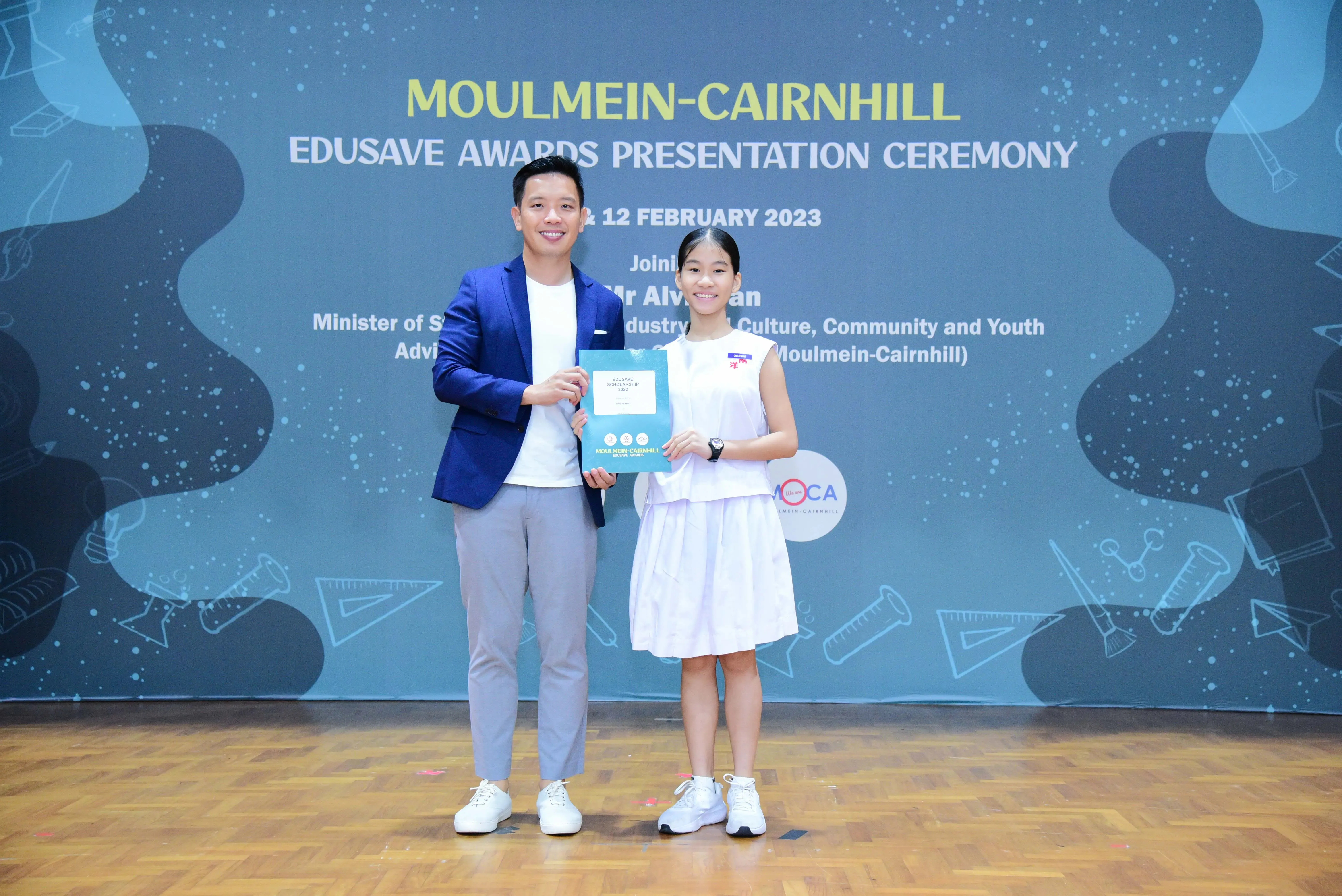 A man in a blue blazer and a young girl in white hold a certificate at the Moulmein-Cairnhill Edusave Awards Ceremony 2023.