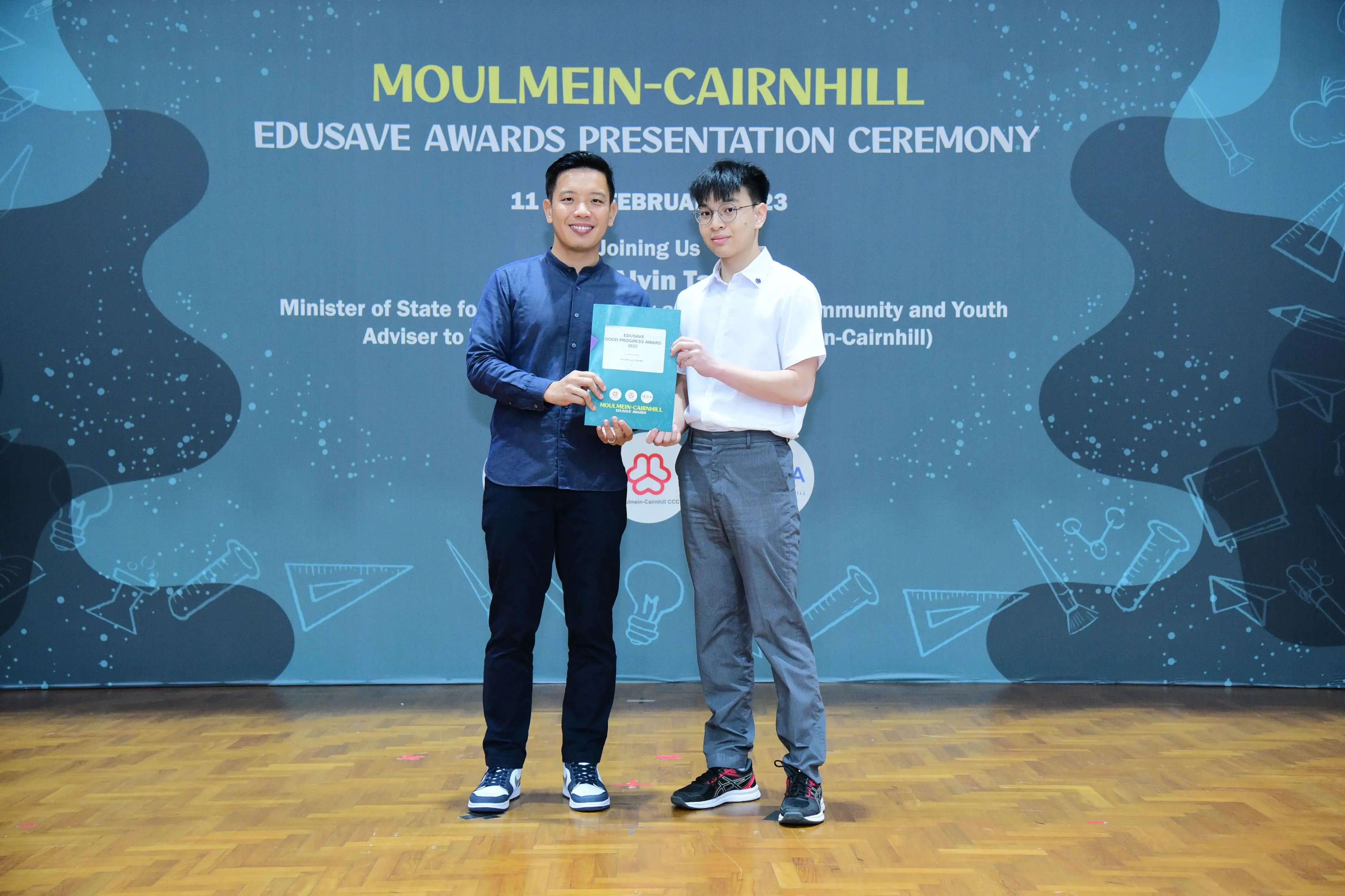 Two men hold an award together at the Moulmein-Cairnhill Edusave Awards Presentation Ceremony backdrop.