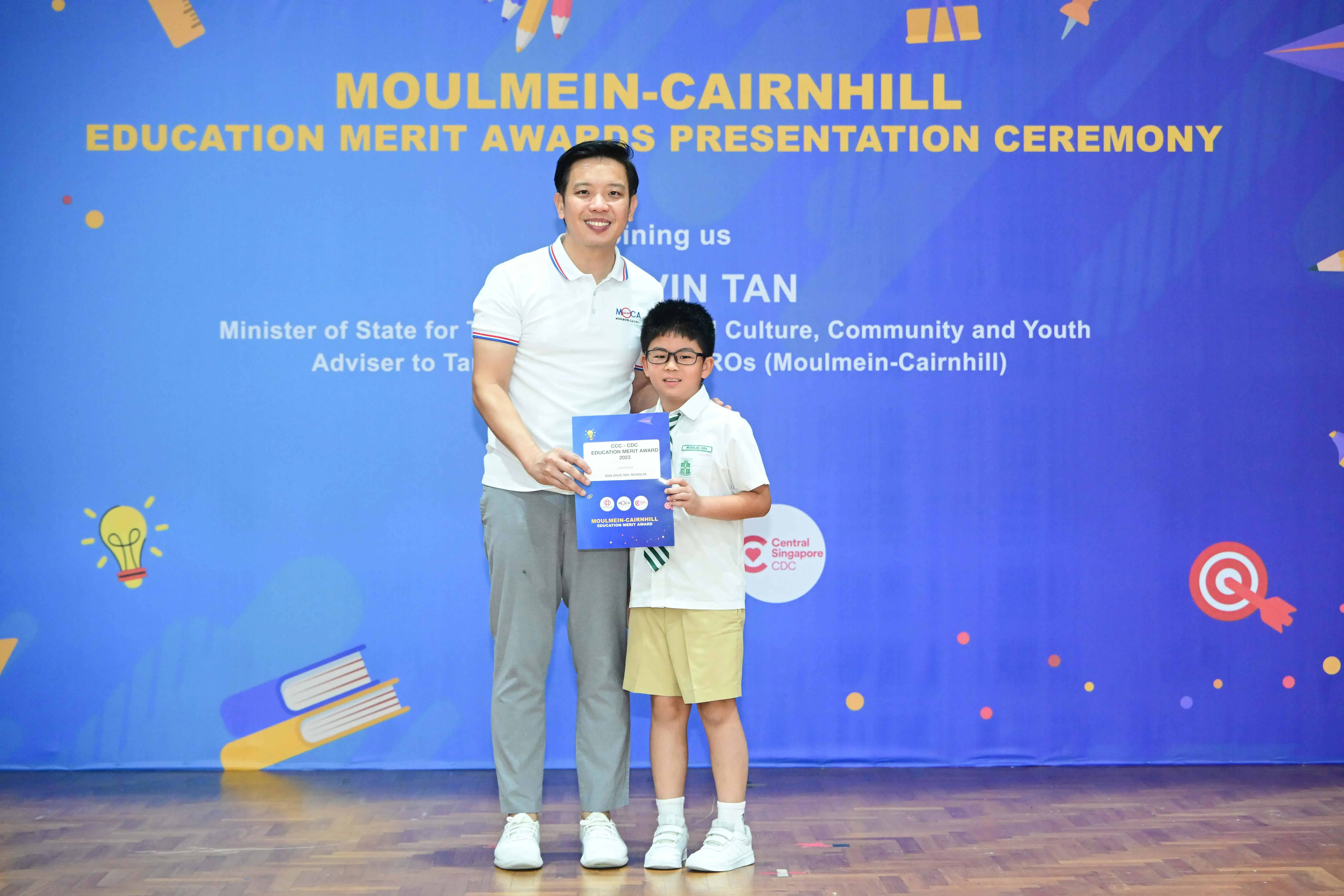 An adult and a young boy in school uniform hold a certificate in front of a Moulmein-Cairnhill Education Merit Awards banner.