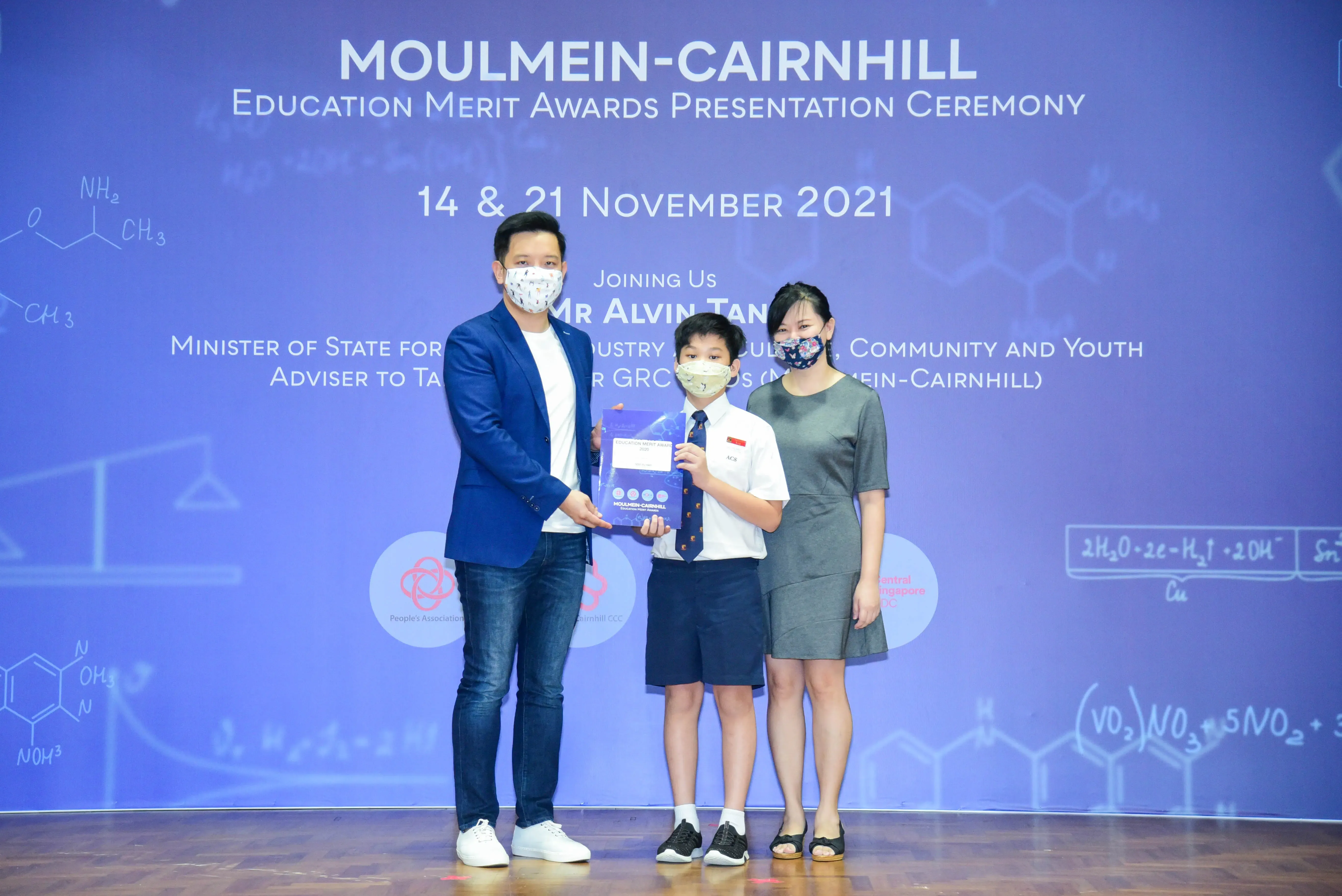 Two adults and a young boy in school uniform hold an award at the Moulmein-Cairnhill Education Merit Awards ceremony.