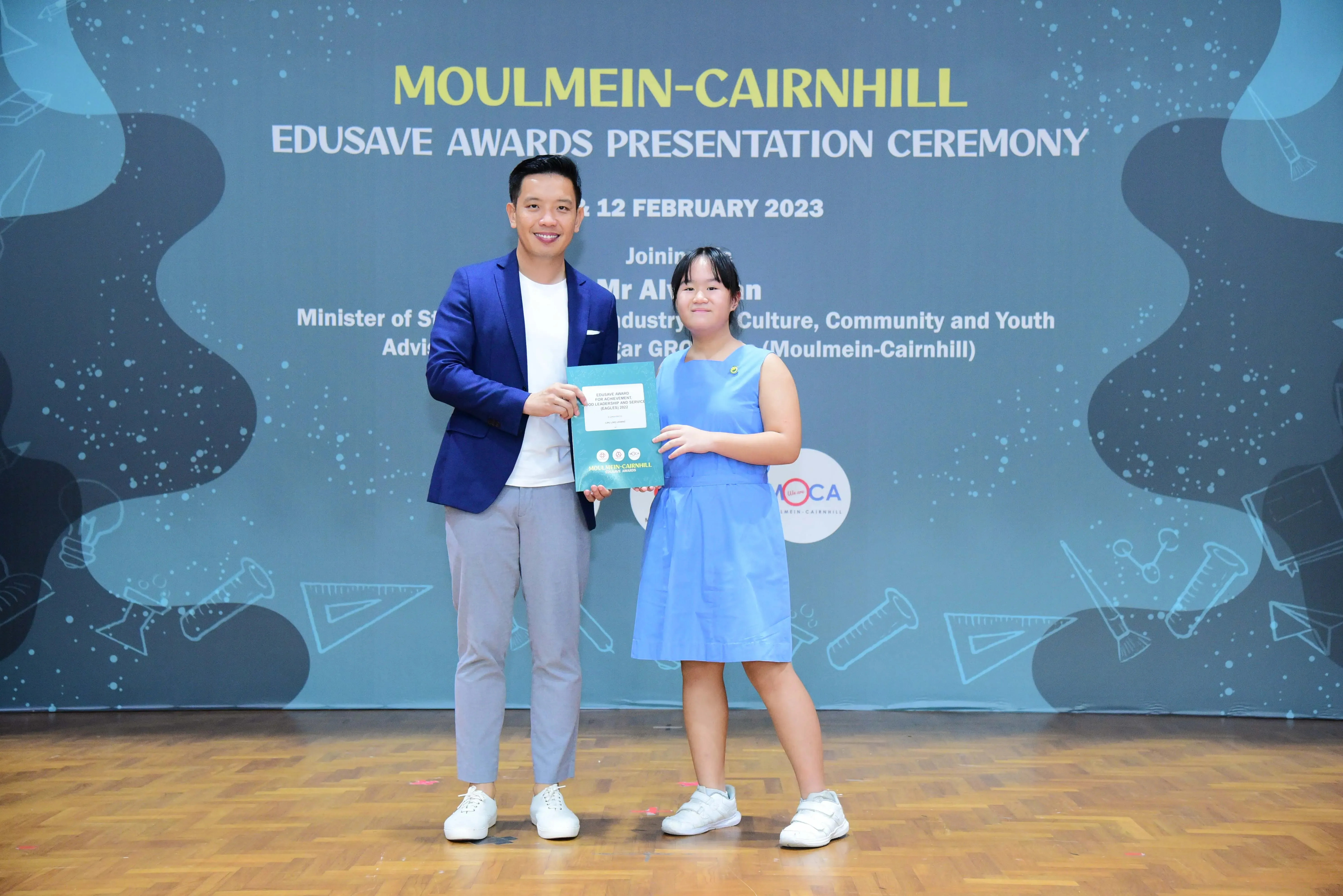 A man in a blue blazer and a girl in a blue dress hold an award together at the Moulmein-Cairnhill Edusave ceremony.