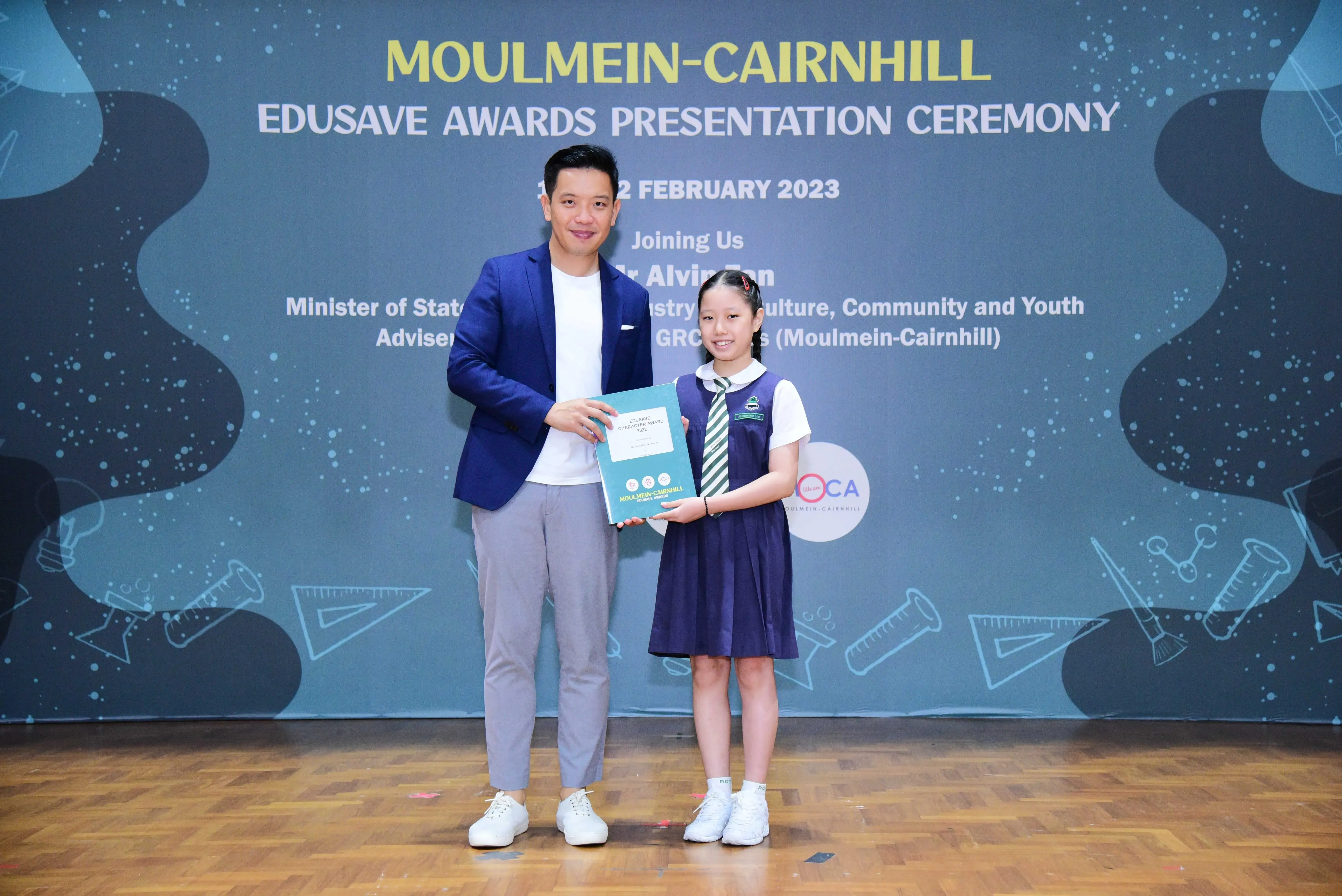 A man in a blue blazer and a young girl in school uniform hold a certificate at the Moulmein-Cairnhill Edusave Awards 2023.