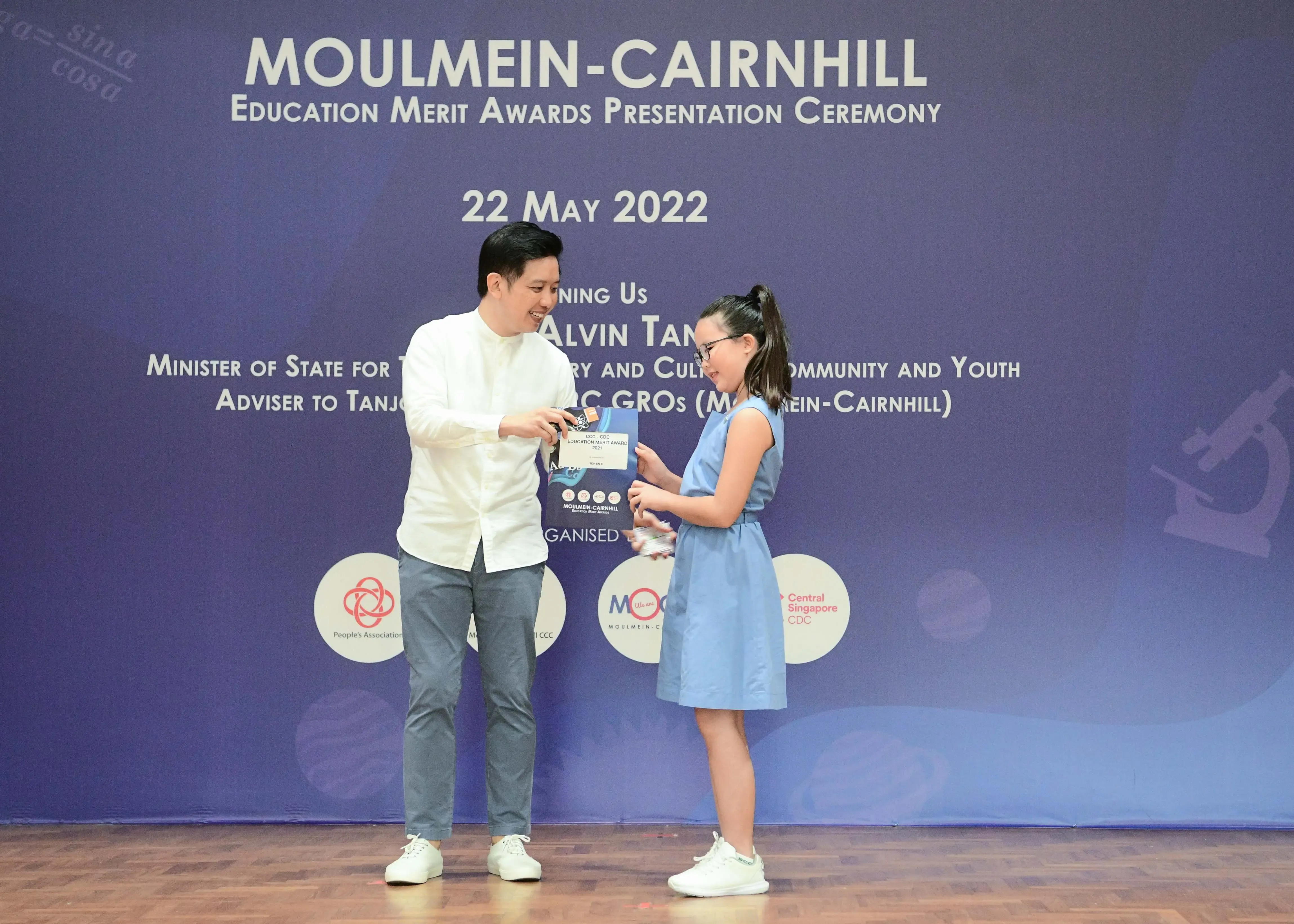 A man in white shirt presents an award to a young girl in blue dress at the Moulmein-Cairnhill Education Merit Awards 2022.
